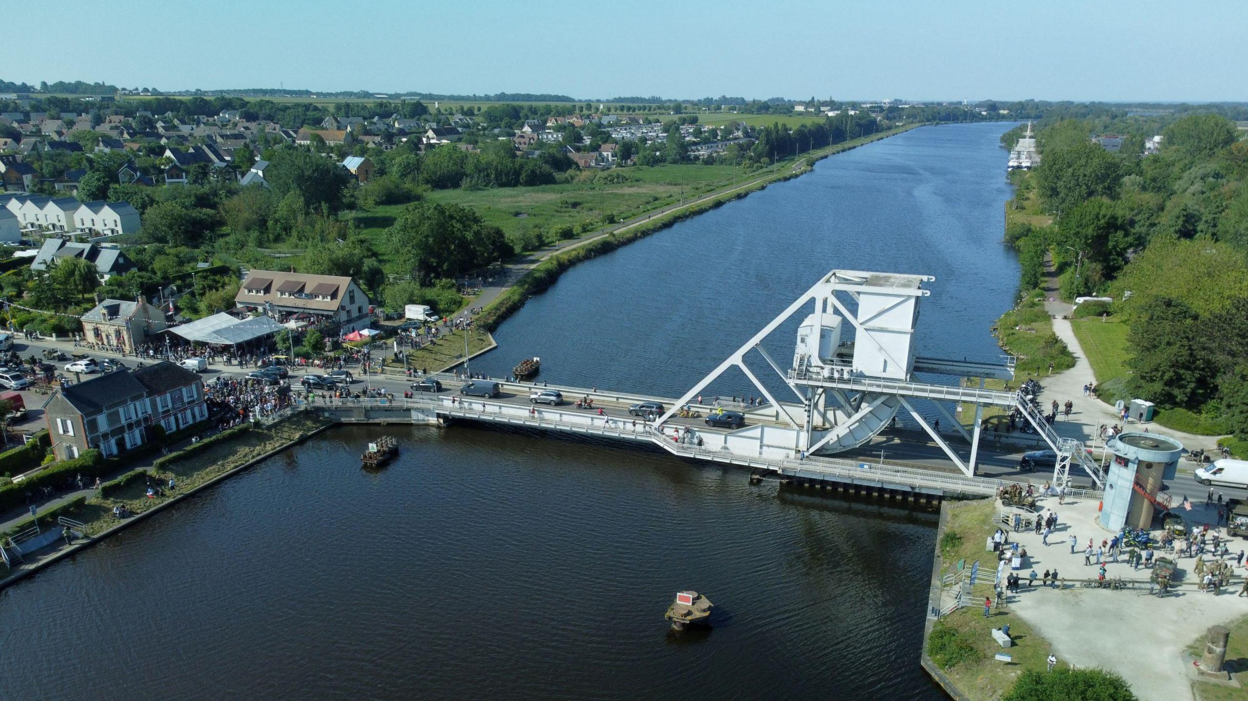 A grey metallic bridge over a wide river. Various military vehicles and personnel can be seen surrounding the bridge