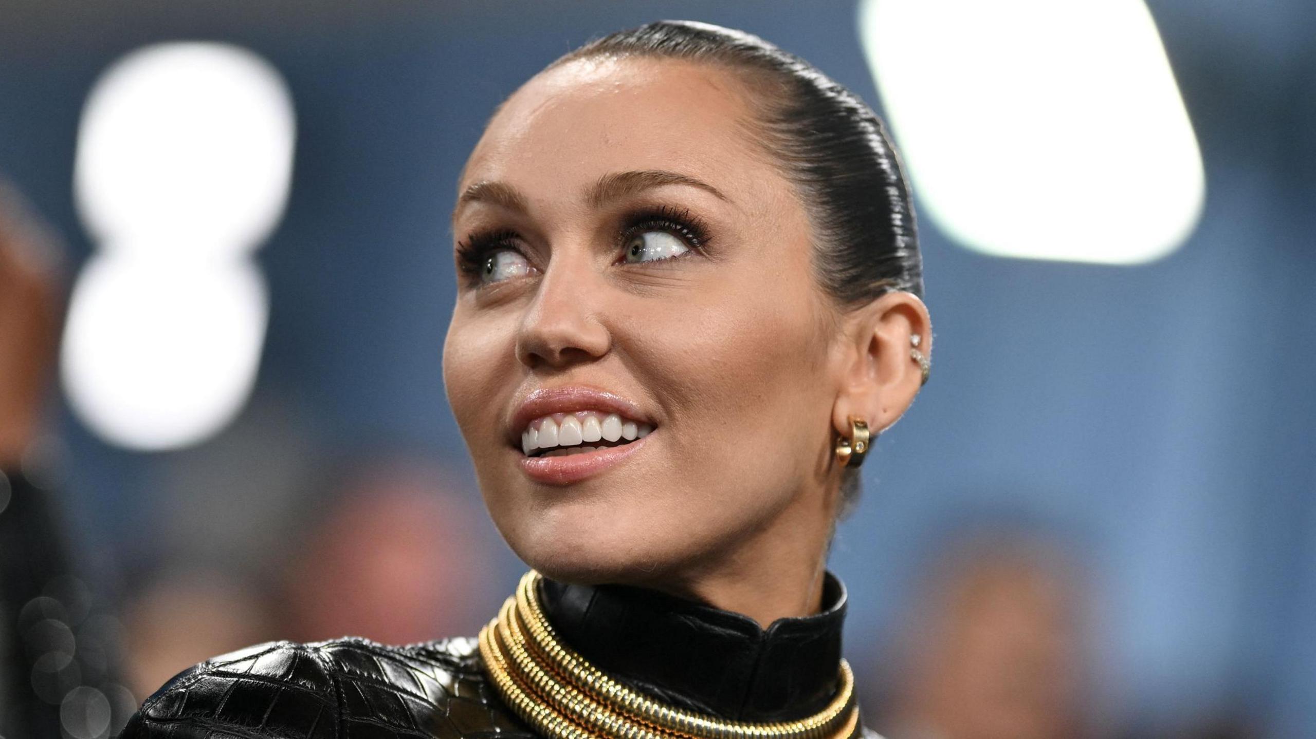 The singer Miley Cyrus is at a glamour music event and is framed by two bright lights that are just out of focus in the background. She has her hair slicked back and is smiling as she looks off into the distance. She is wearing a shiny black outfit with gold trim on the neck.