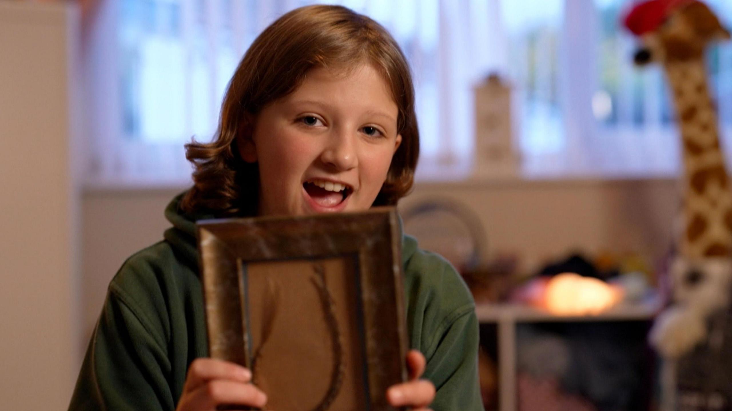 Llun o Kathryn yn gwenu ar y camera. Mae'n gwisgo siwmper du ac mae ganddi wallt brown. Mae'n dal ffrâm gyda darn o'i gwallt ynddi.