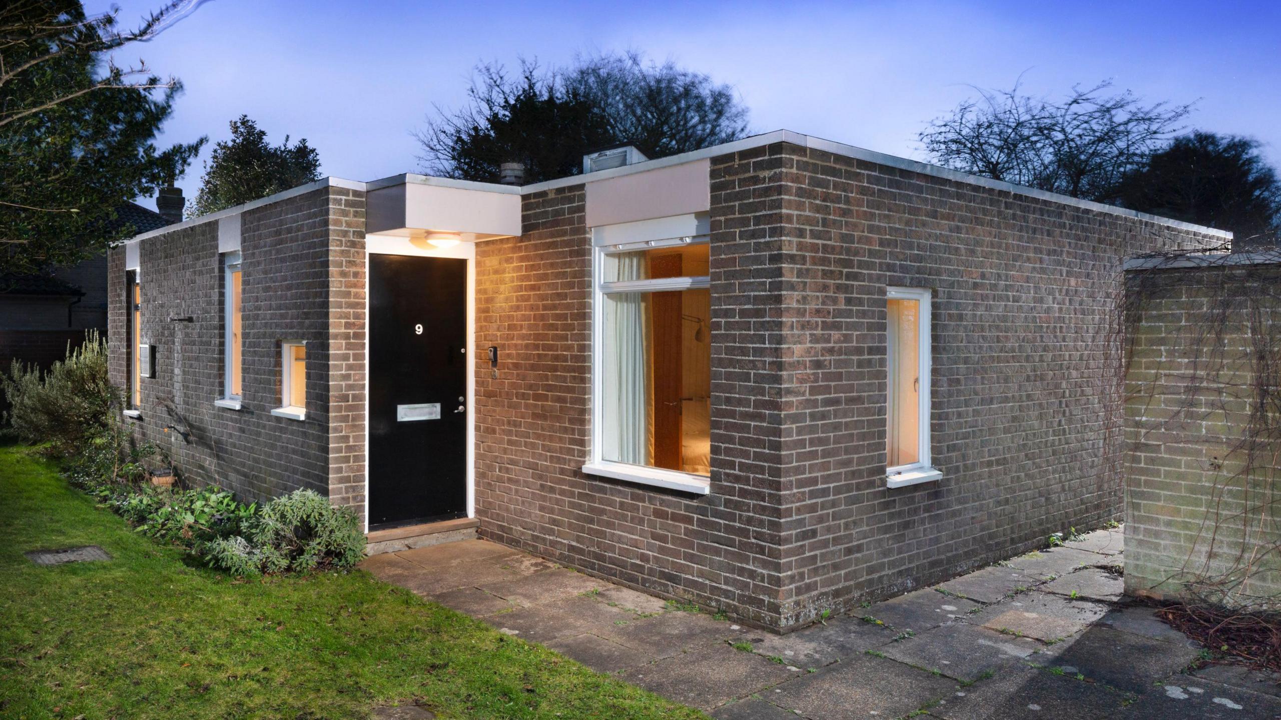 A view of the exterior of 9 Church Walk in Aldeburgh. It is a red brick bungalow which has faded. It has a black door and windows with lights on inside.