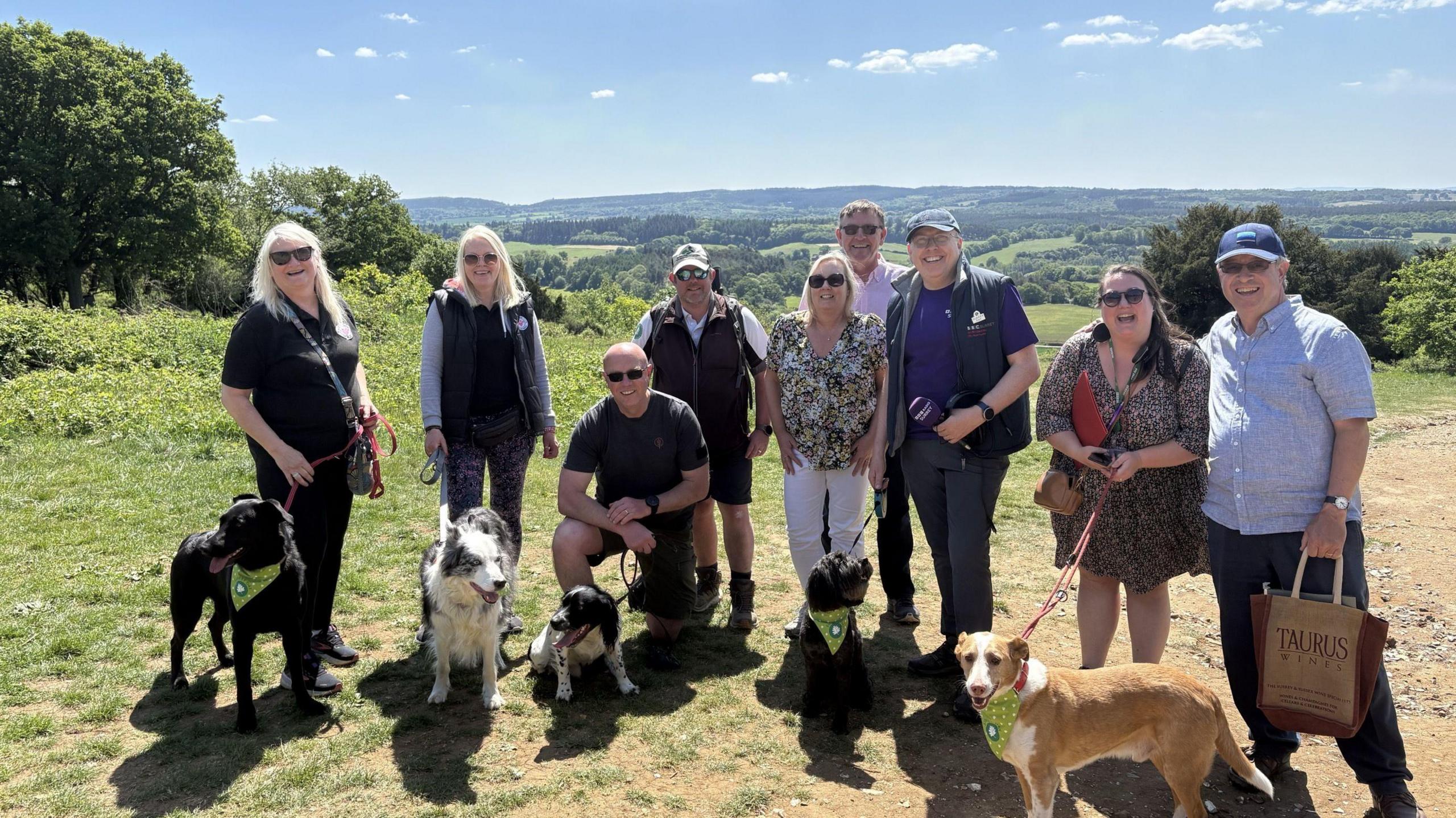 A group of nine people stood in the countryside on a sunny day, with a green vista behind them. One man is carrying a microphone and there are five dogs on leads.