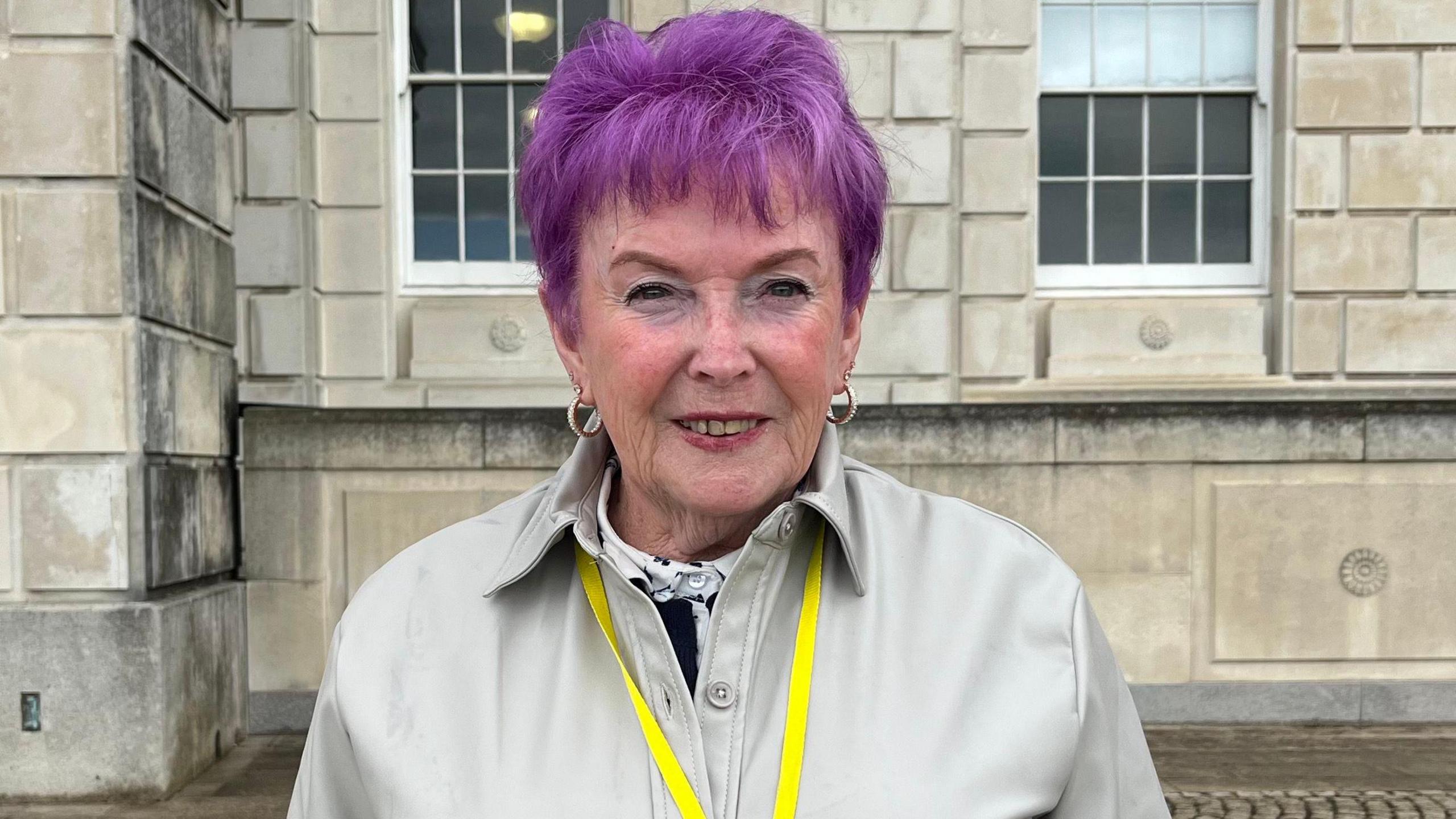 A pistillate with purple hairsbreadth and tan overgarment smiling astatine the camera, she has a yellowish lanyard