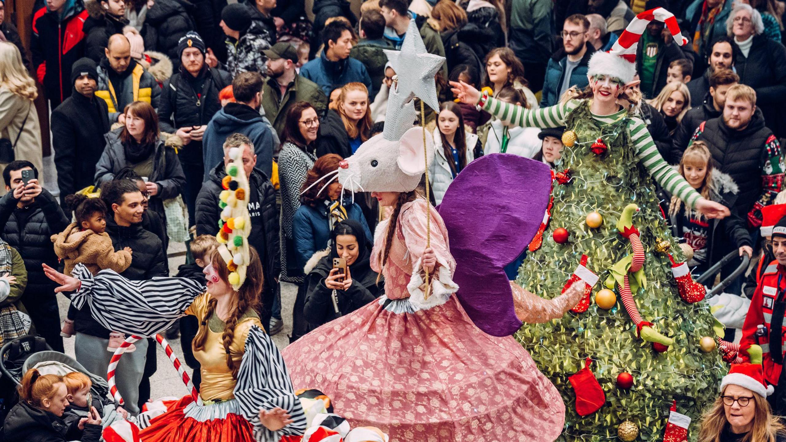Artists dressed up as Christmas characters including a Christmas tree for a  Christmas parade among hundreds of people smiling. 