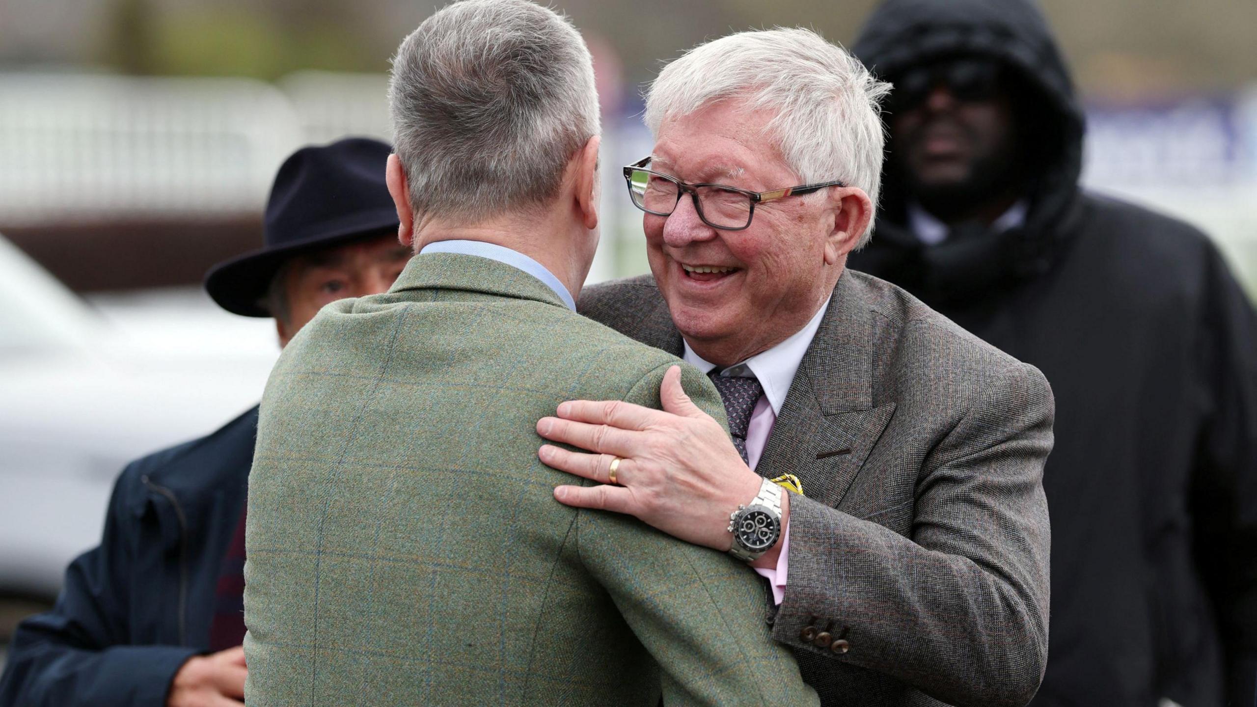 Former Manchester United manager Sir Alex Ferguson, wearing a grey suit, smiles as he embraces a man, who is facing away from the camera, on day three of the Cheltenham Festival