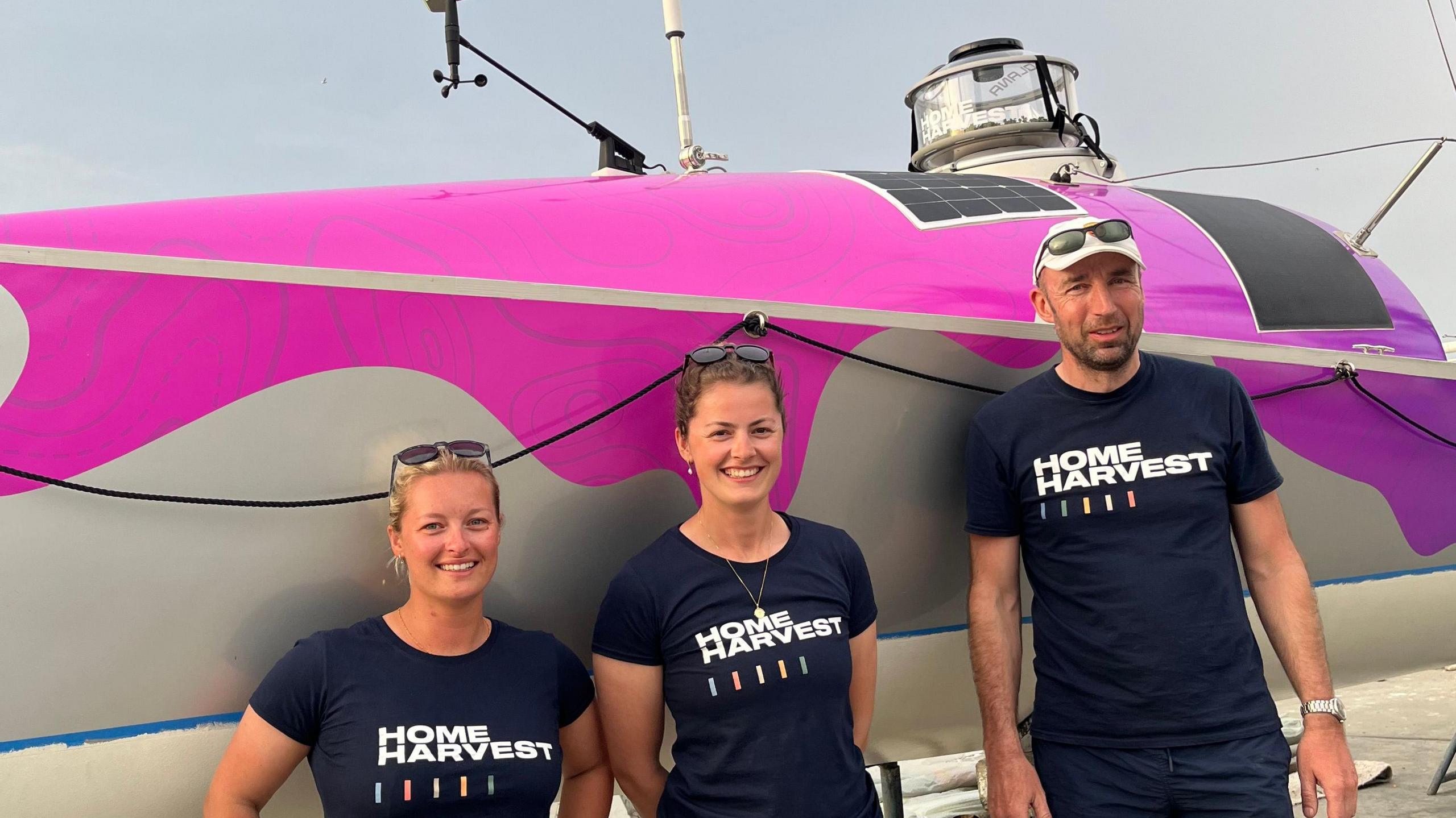 Jess Rowe, Miriam Payne and Andrew Johnson stand in front of a rowing boat. They are all wearing t-shirts saying 'Home Harvest'. On top of the boat is the "smart farm" device.