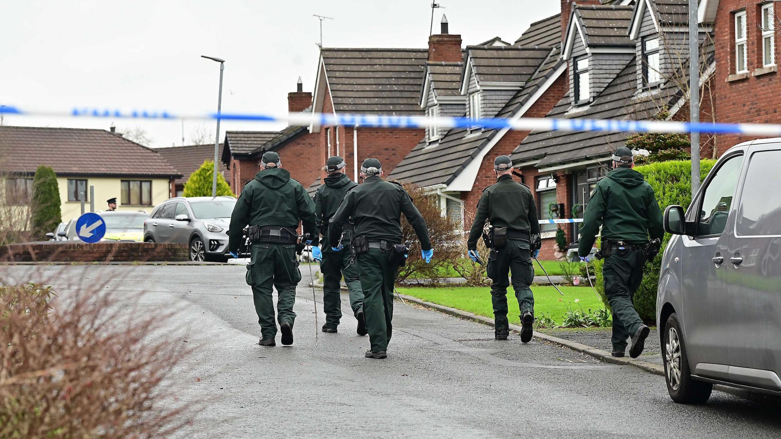 Five police officers wear green and black uniforms with black hats. They are walking up a concrete residential street with red brick houses on their right hand side, they have their backs to the camera. 