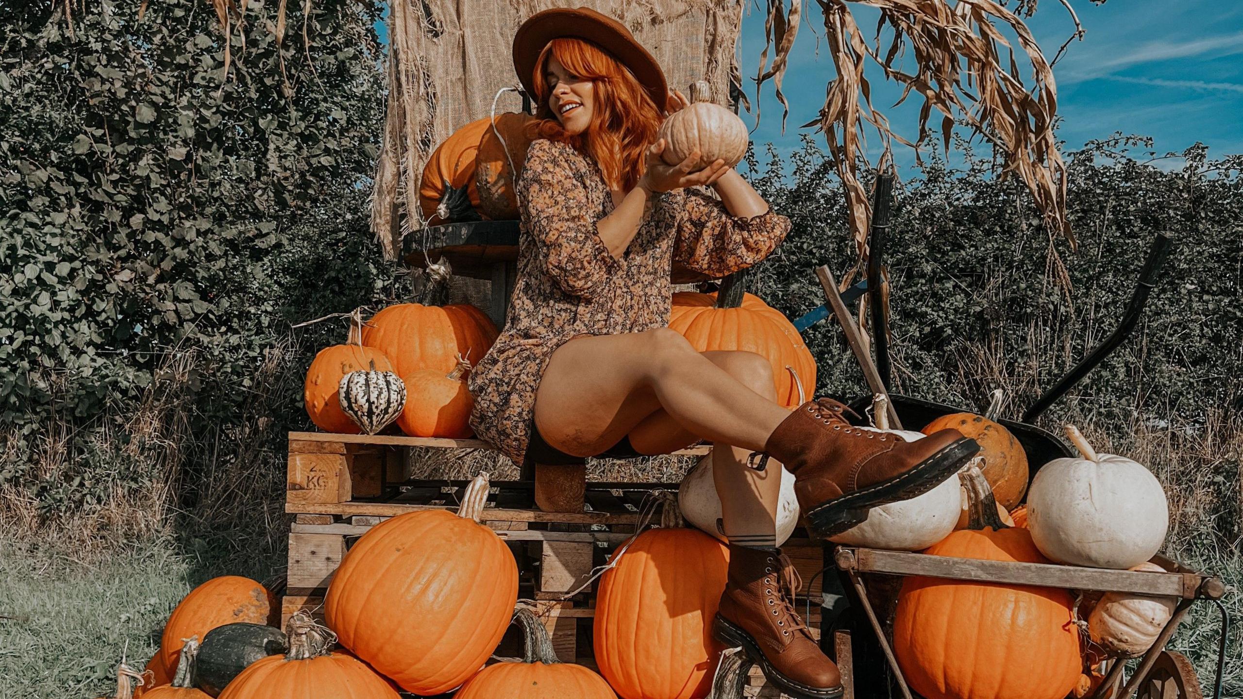 A woman with red hair sits on top of a wooden crate, surrounded by pumpkins. She is wearing a hat, red boots and a dark patterned sun dress