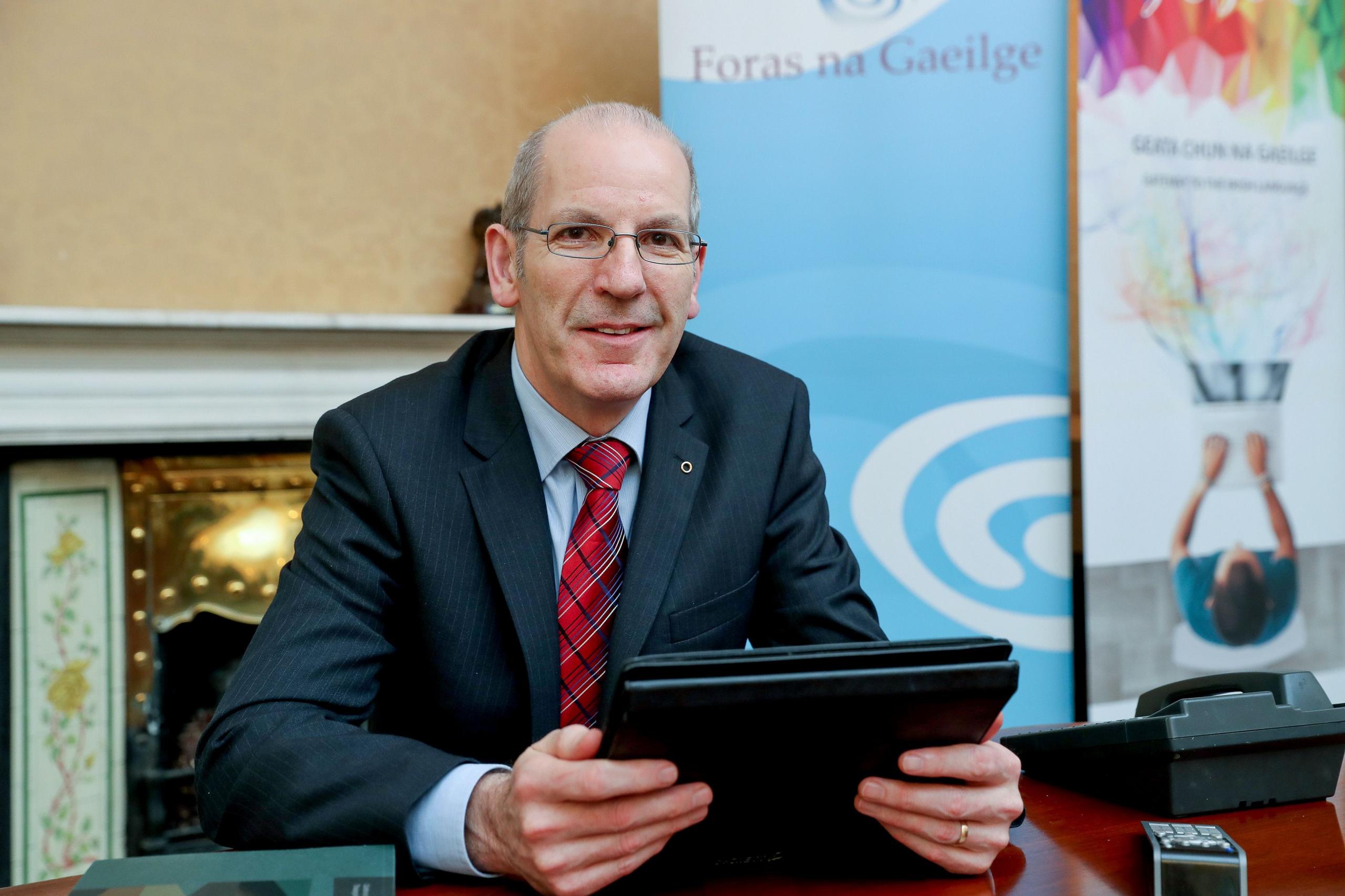 Seán Ó Coinn in a black suit, blue shirt and red checked tie, holding a tablet in an office with the Foras na Gaeilge backdrop beside a mantlepiece.