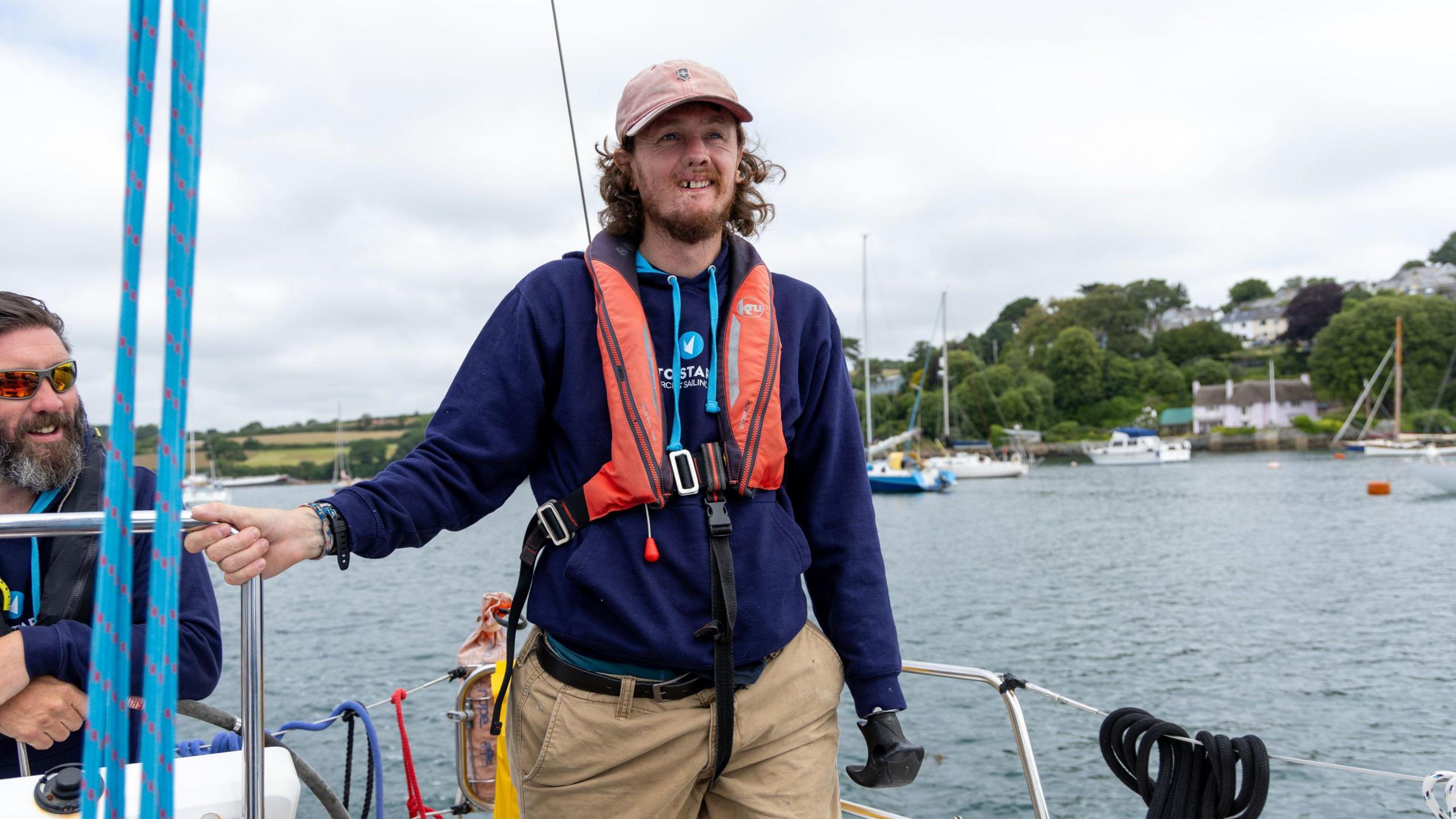 A man in his thirties standing on a boat. He is wearing a pink cap, a life jacket and beige shorts. A man with sunglasses and a beard sits alongside him.