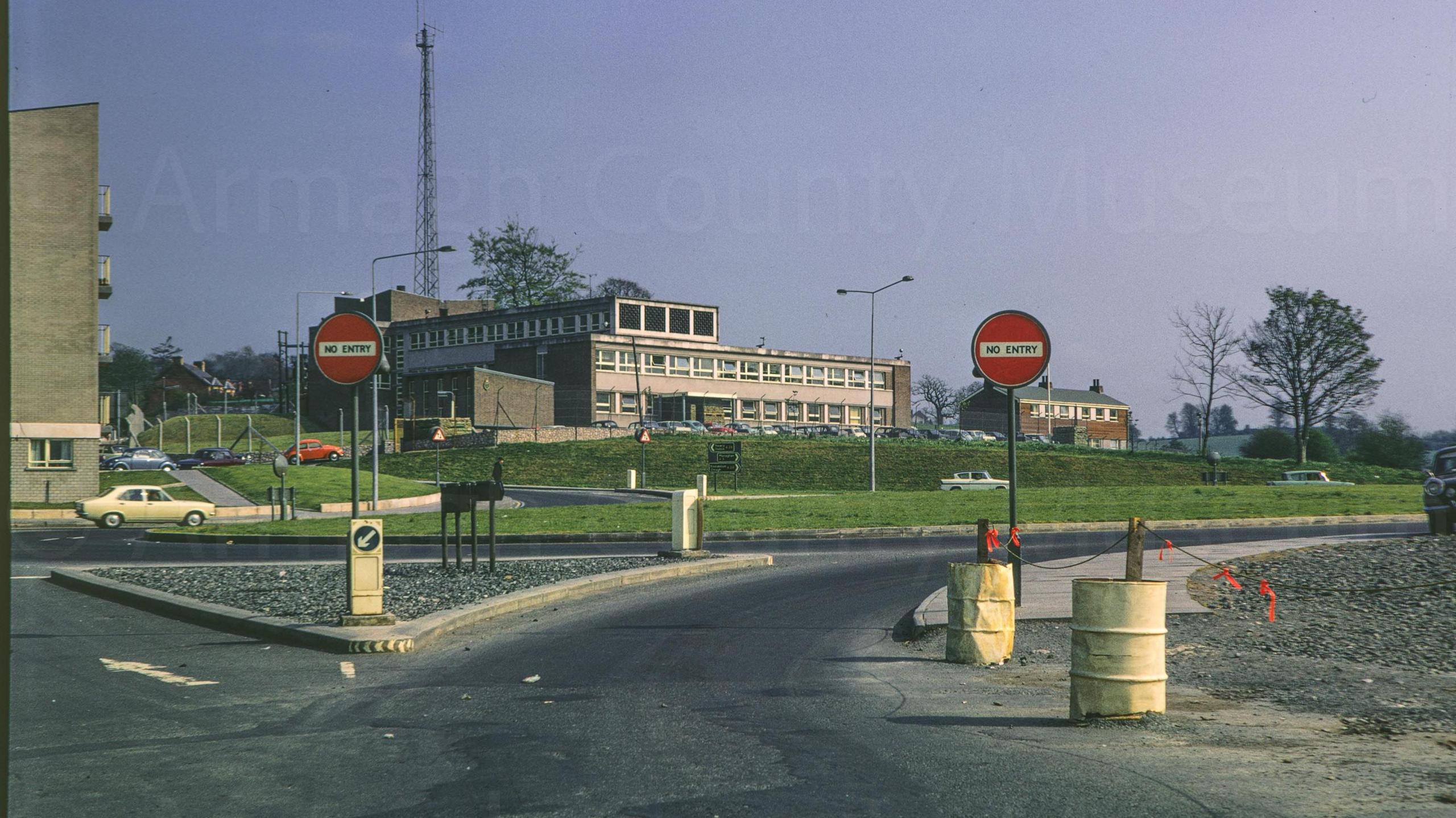 A digitalised photo from a film of abuilding with a lot of windows. It has a road with no entry signs leading up to it. Looking south from junction of Scotch Street and Barrack Street with Newry Road police station in centre frame