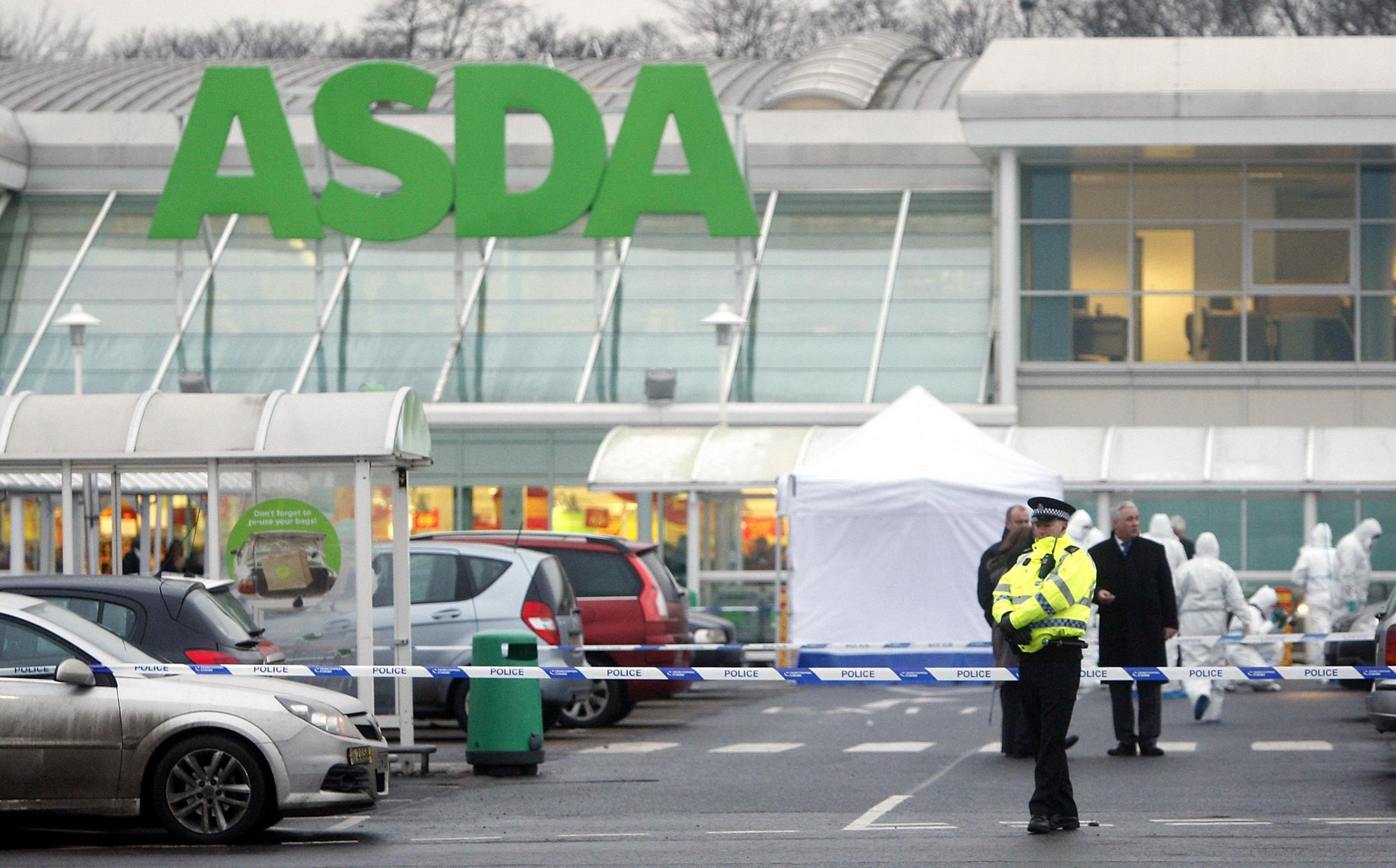 A lone police officer stands guard in front of blue and white police tape in the car park of Asda, Robroyston. Behind the tape a white forensic tent has been erected and specialists in white hazmat suits can be seen carrying out inquiries. Plain clothes detectives, including one wearing black trousers and a long black coat, are also visible in the background.