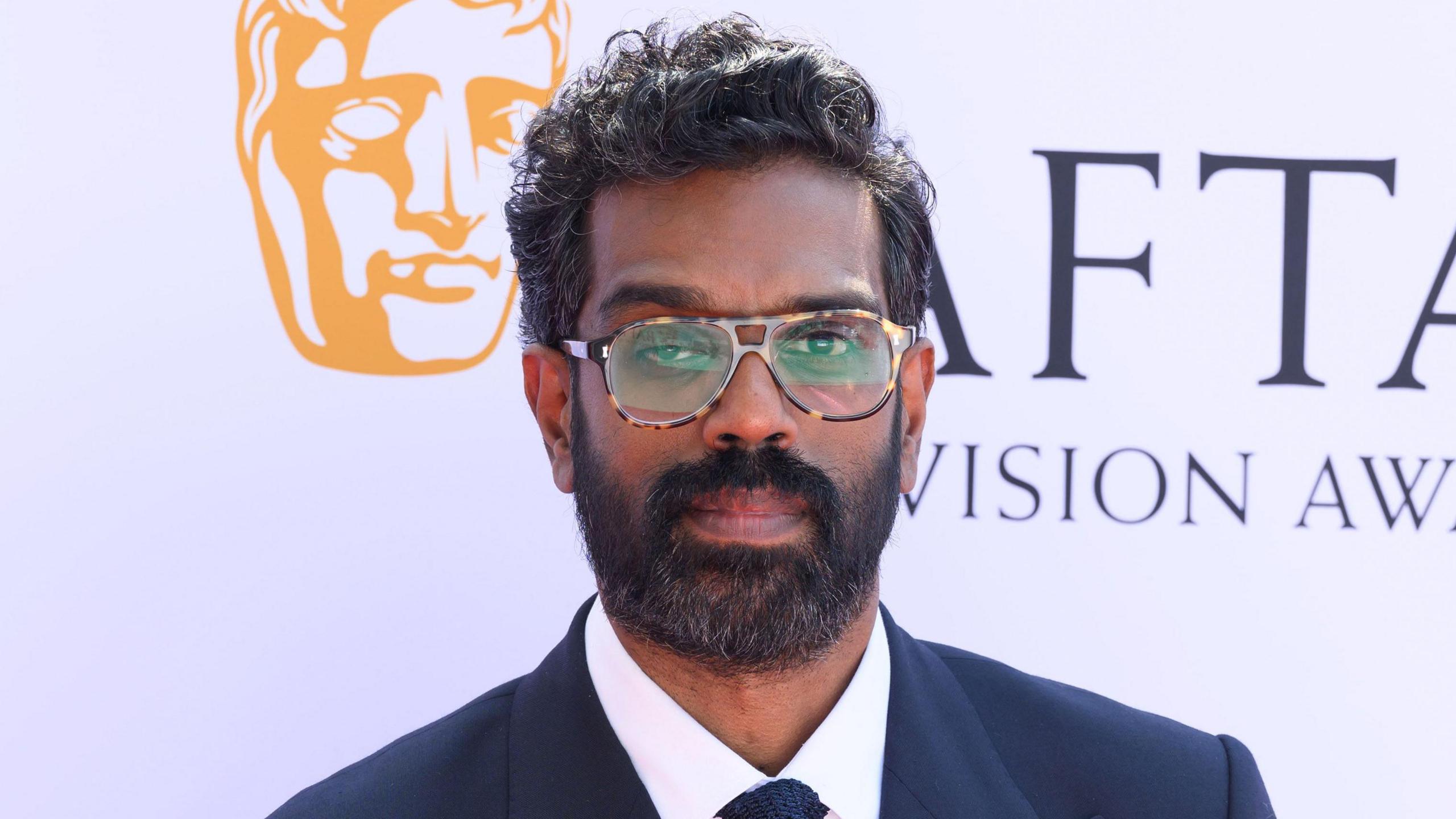 Romesh Ranganathan in a suit standing in front of a white backdrop.