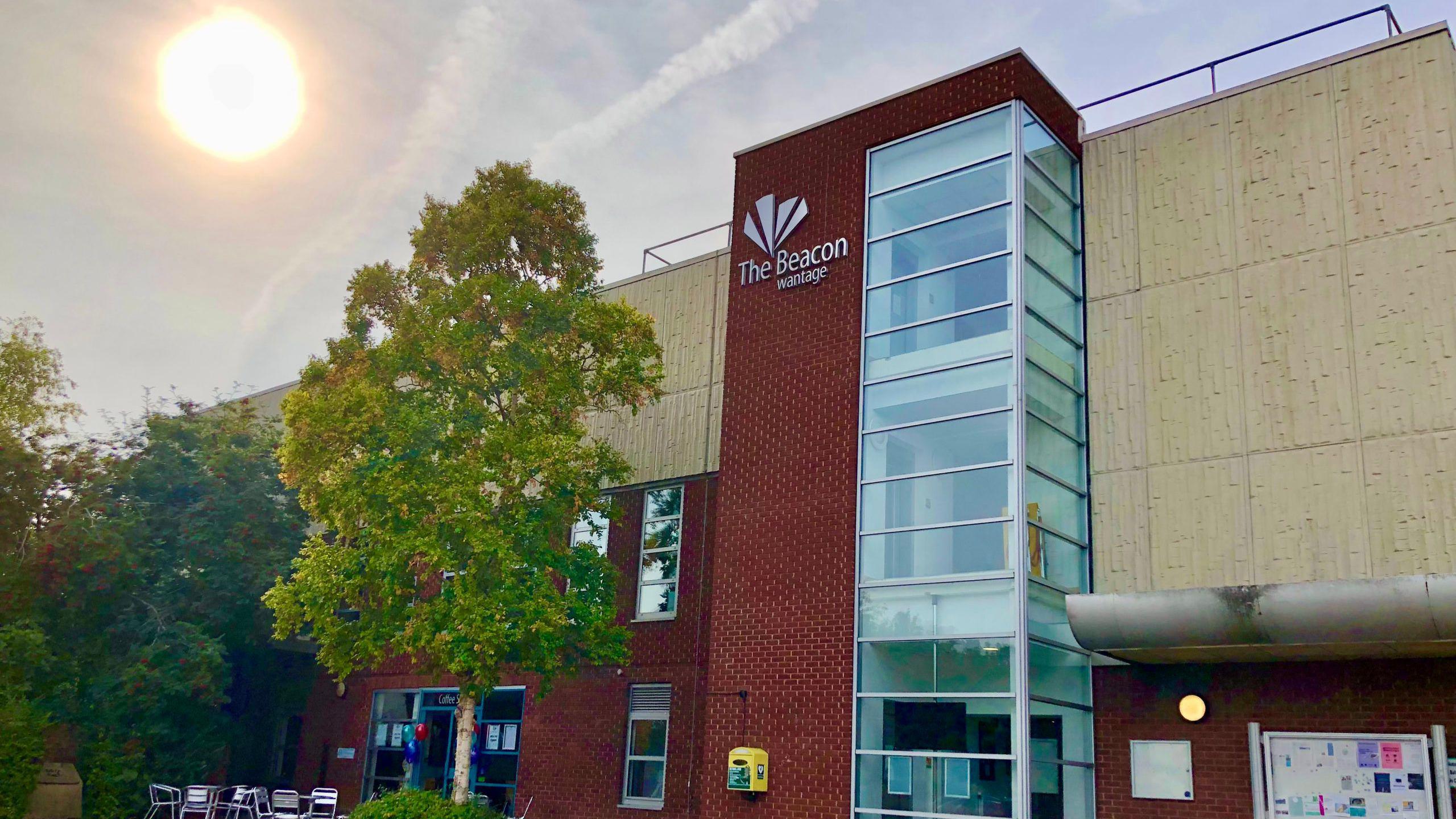 A picture of The Beacon Arts Centre in Wantage, from outside the front entrance. It is a mostly red brick building, with The Beacon logo on a central pillar and several trees out the front.