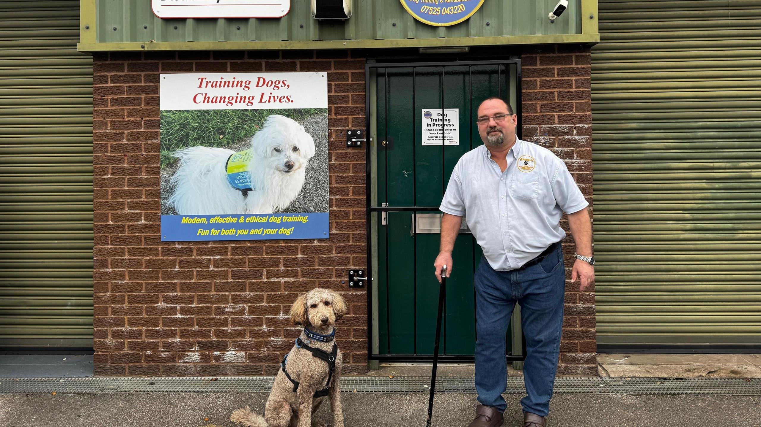 A man with short dark hair and dark facial hair is wearing glasses, a white shirt and dark blue jeans. He is holding a black walking stick and is standing next to a beige dog with short curly hair. They are standing outside a red brick industrial unit building with green corrugated metal doors. A sign with a white fluffy dog on it reads "training dogs, changing lives"