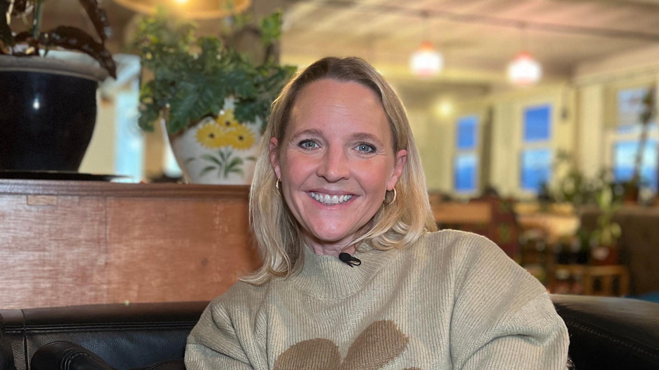 Kate Wright looking at the camera. She is smiling and wearing a light grey jumper. She is sat down and has a wooden shelf behind her.