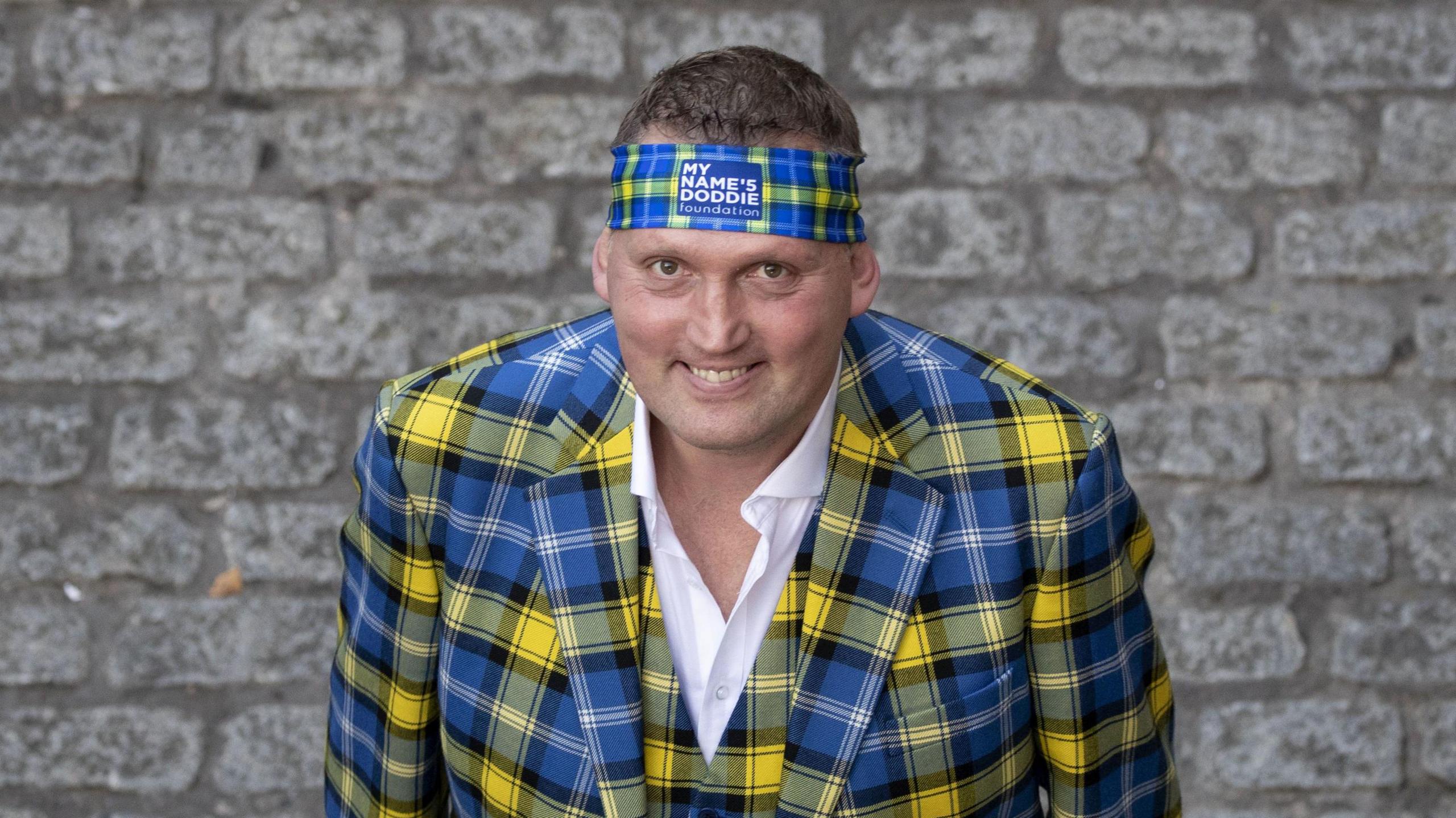 Doddie Weir is wearing a yellow and blue tartan three piece suit and headband.