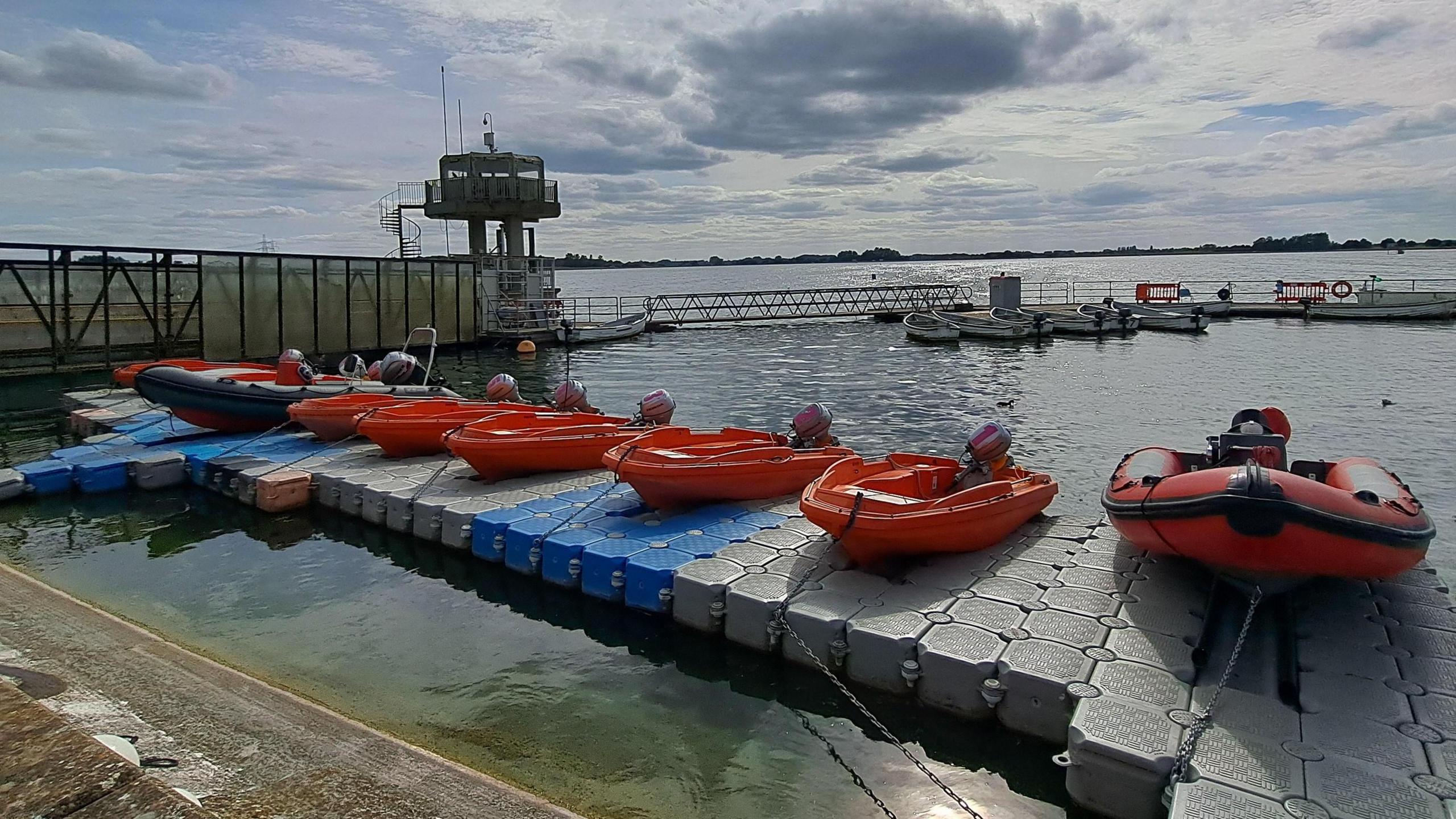 A cloudy bright sky with blue patches gives way to shimmering water of a reservoir. A works tower is to the right of the image in the water, in front is a pontoon with several orange and blue boats each with outboard motors at their rear. Set on the water behind them are a few small white boats floating on the water.