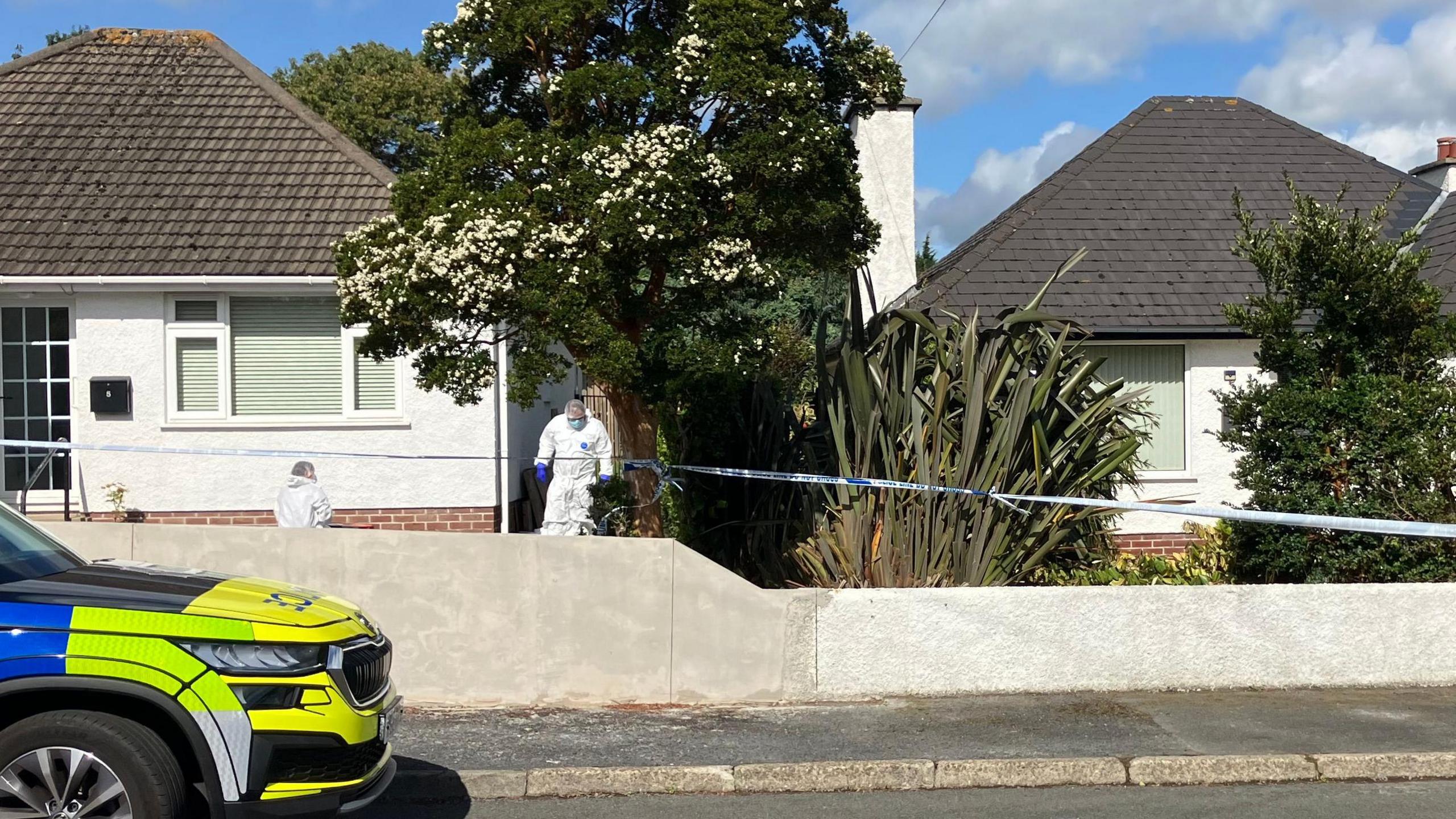 Two white houses with brown roofs are surrounding with blue and white police tape as forensics in white suits examine the exterior. There is a blue, yellow and grey police car park on the street in front of the house.