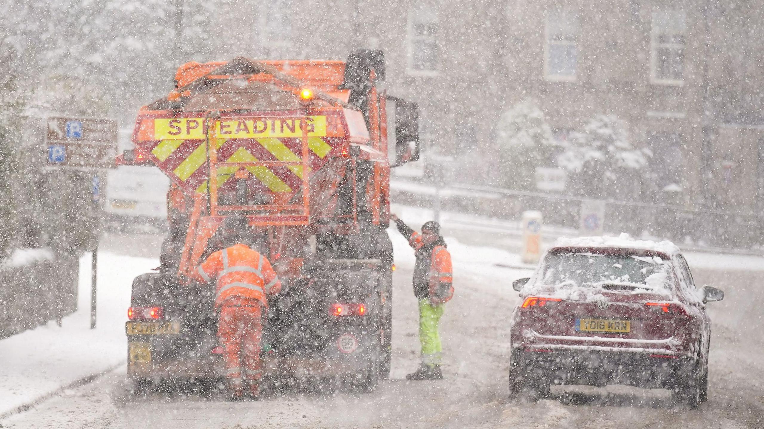 A gritter stopped on the side of the road in the snow with two workers in hi-vis around the vehicle. There is a dark red car passing by the side of the gritter.