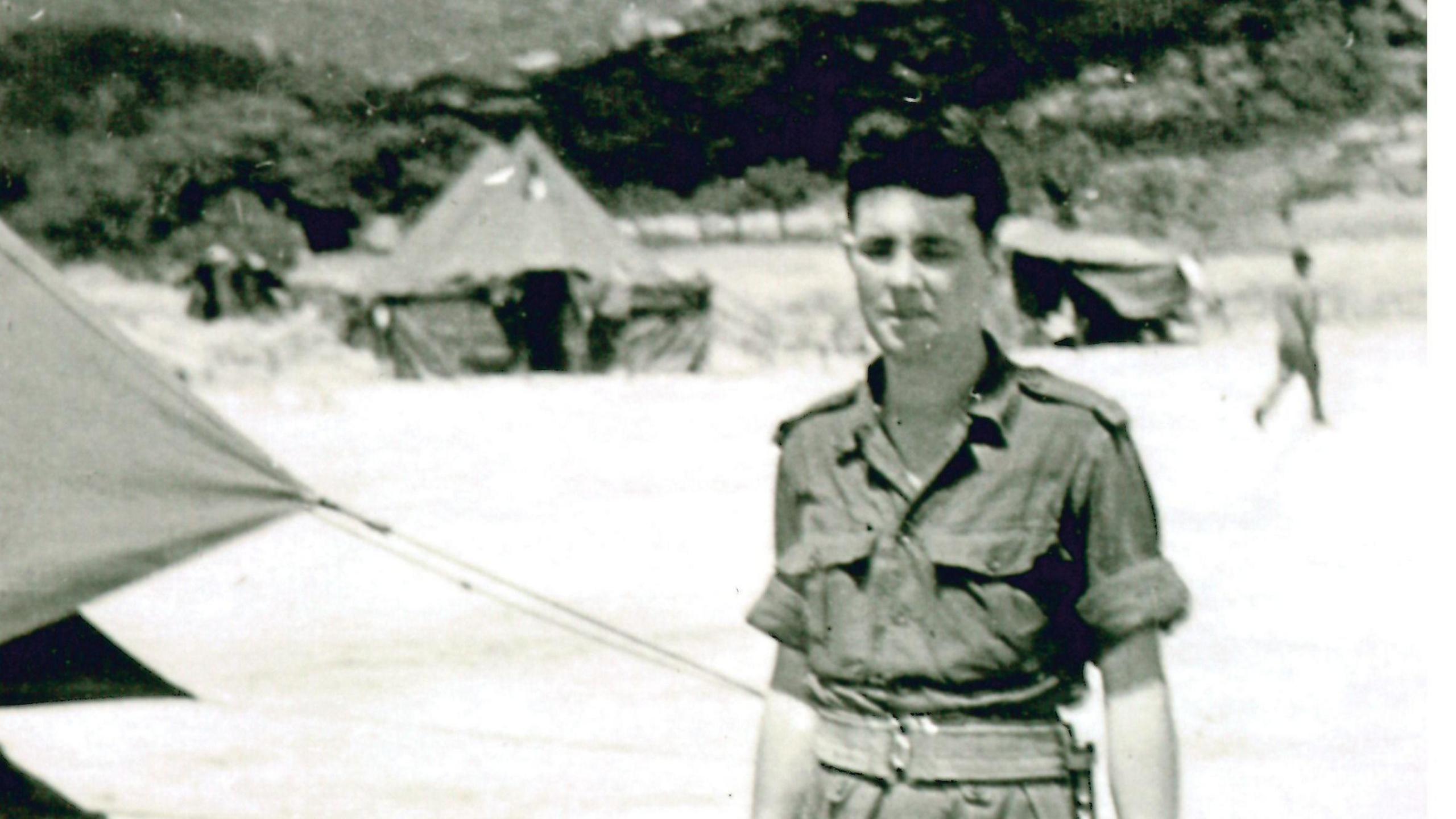 Albert, with thick black hair wearing uniform, stands amidst a camp of tents