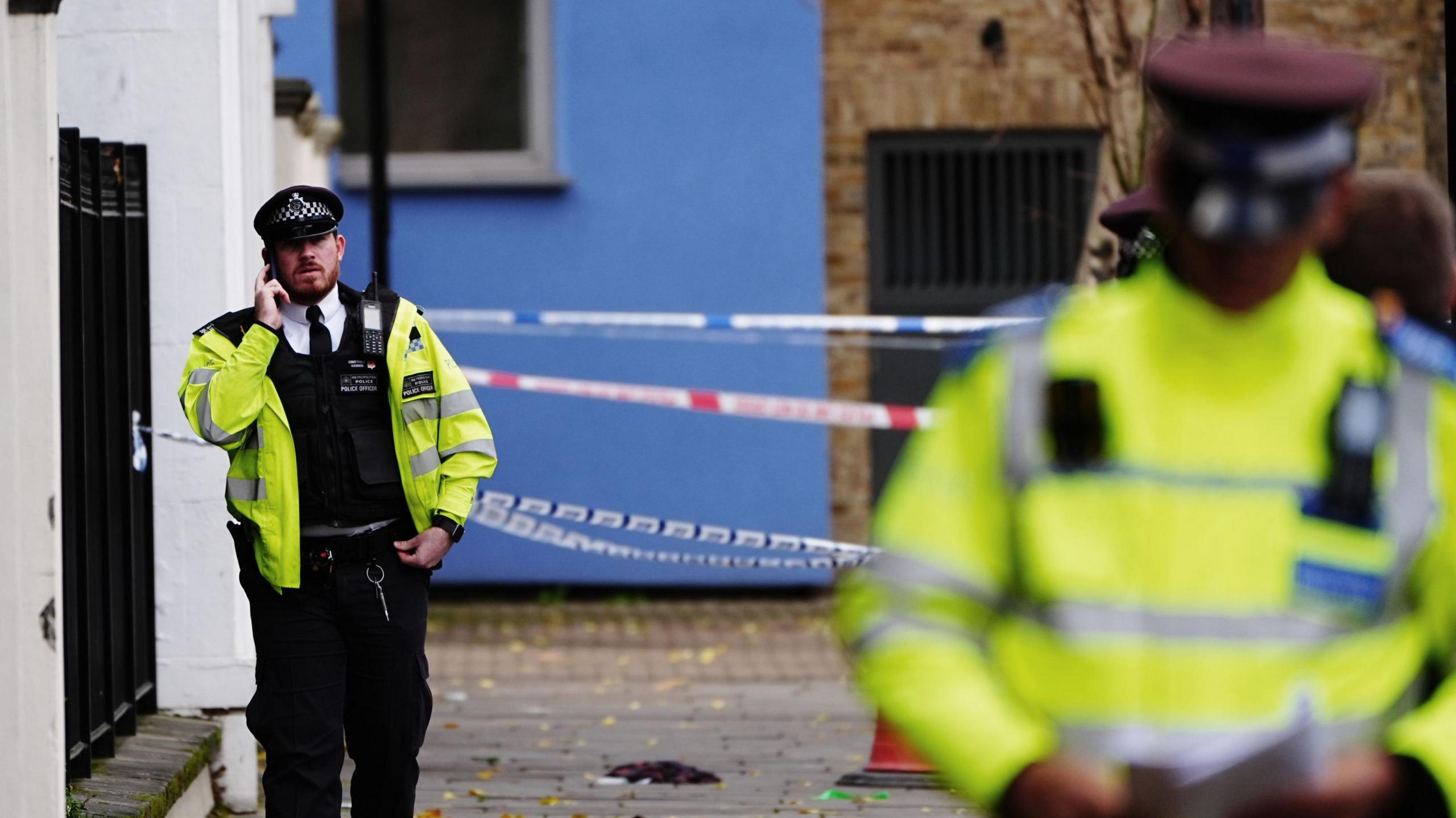 A uniformed police officer uses a mobile phone while walking past a cordon on part of the street. Several out-of-focus police officers stand in uniform on the right of the image