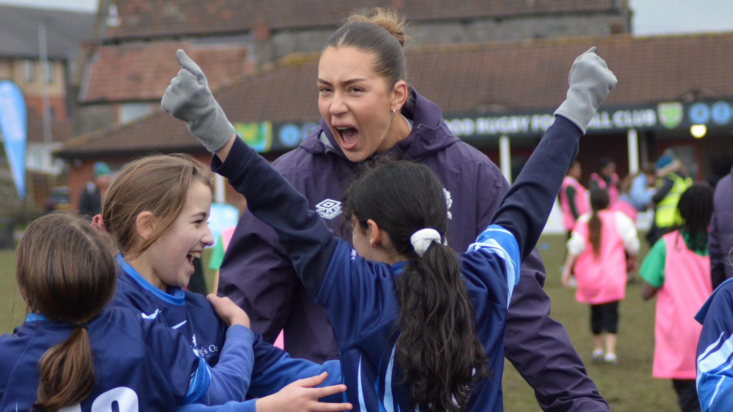 An adult female rugby player in a blue kit cheers with her mouth wide open as she celebrates with younger players, also in blue, as part of a training session