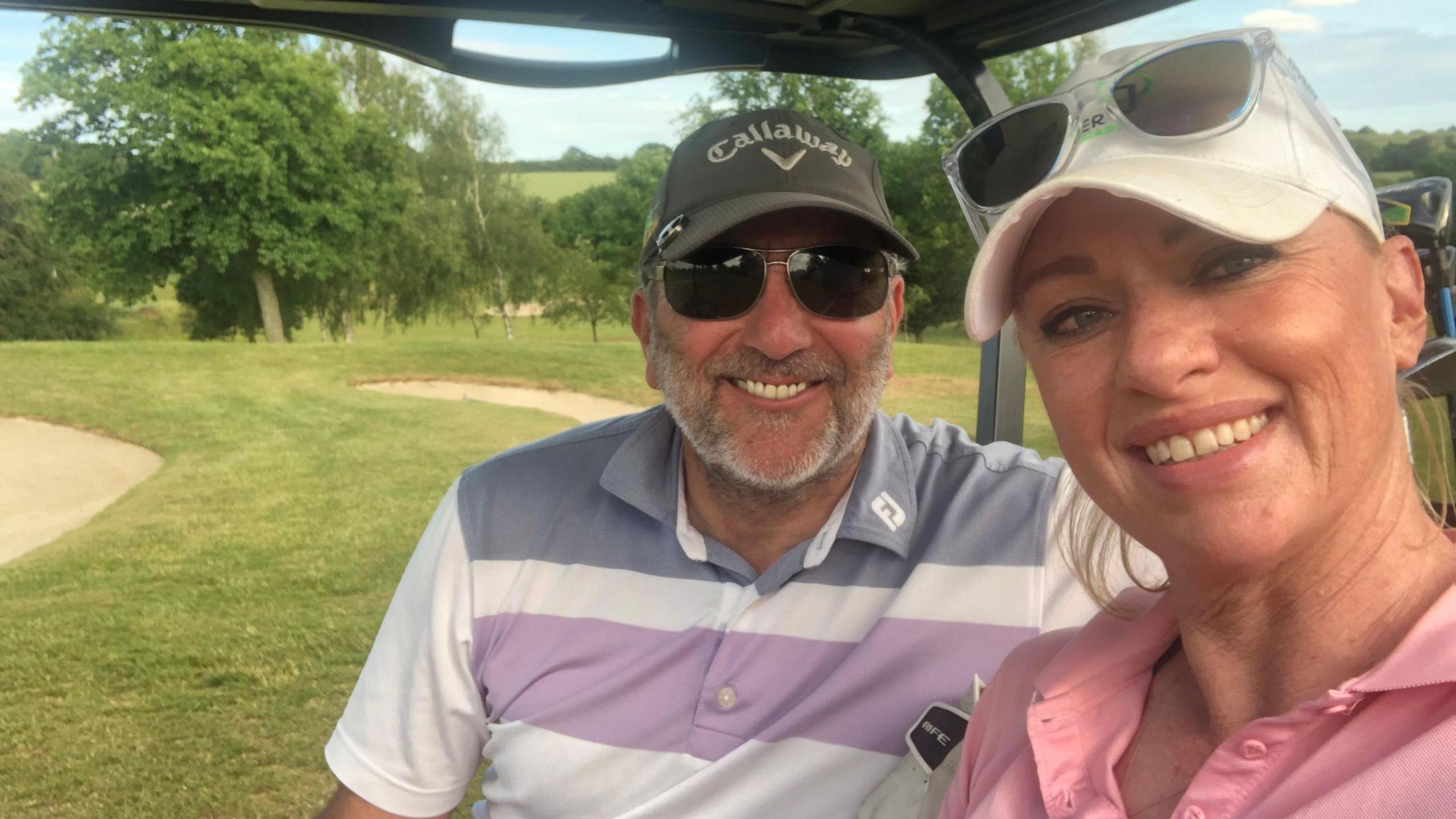 Joanne Oliver and Robert in a golf buggy on a golf course. Joanne is to the right wearing a white cap, with sunglasses on top and wearing a pink polo shirt, Robert is smiling straight at the camera, wearing a stripped polo shirt, with a cap and sun glasses on. 