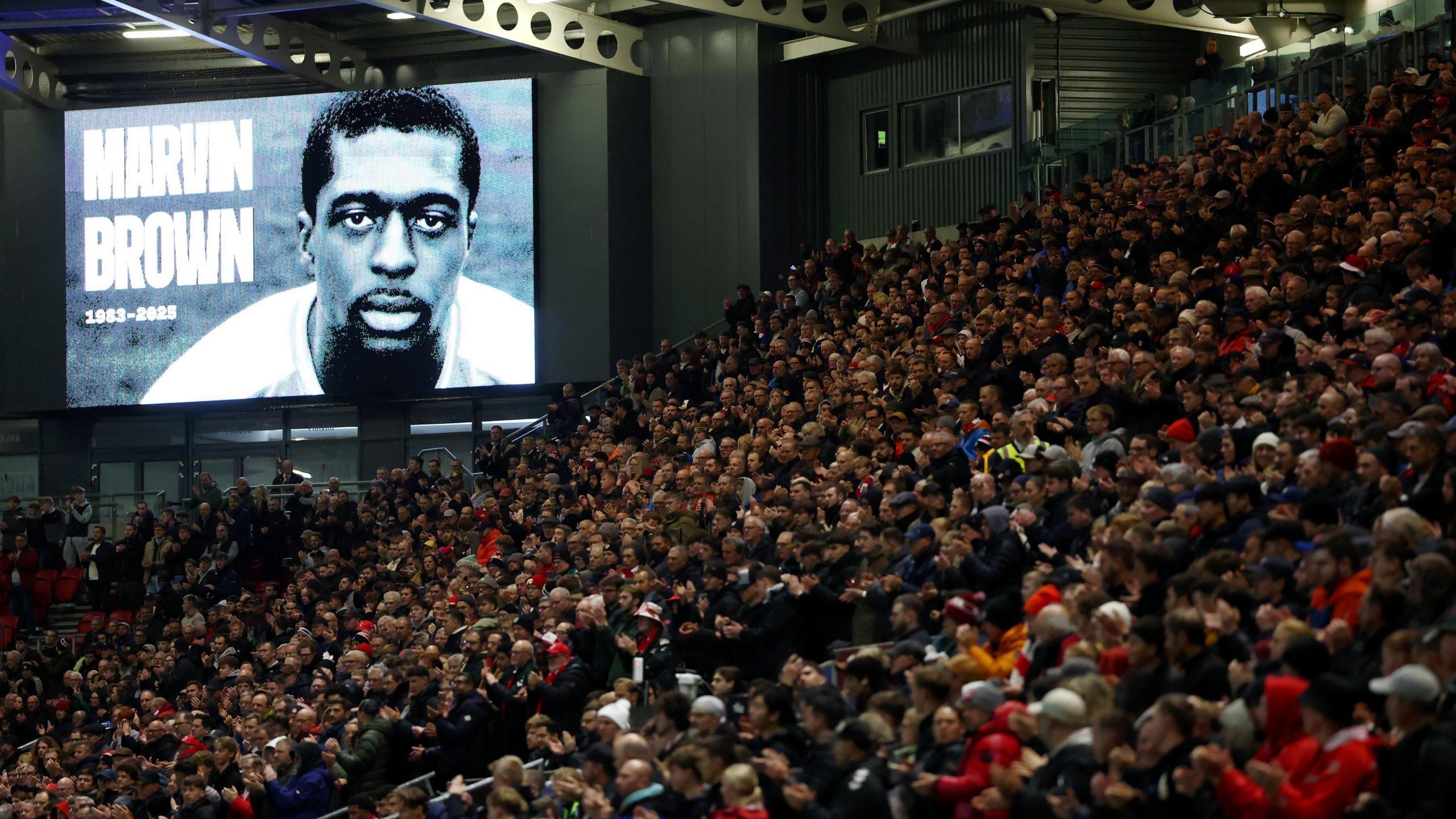 Hundreds of Bristol City fans are seen standing in one of the stands at Ashton Gate during the match with Blackburn Rovers. On a large screen above them is a black-and-white image of former player Marvin Brown.