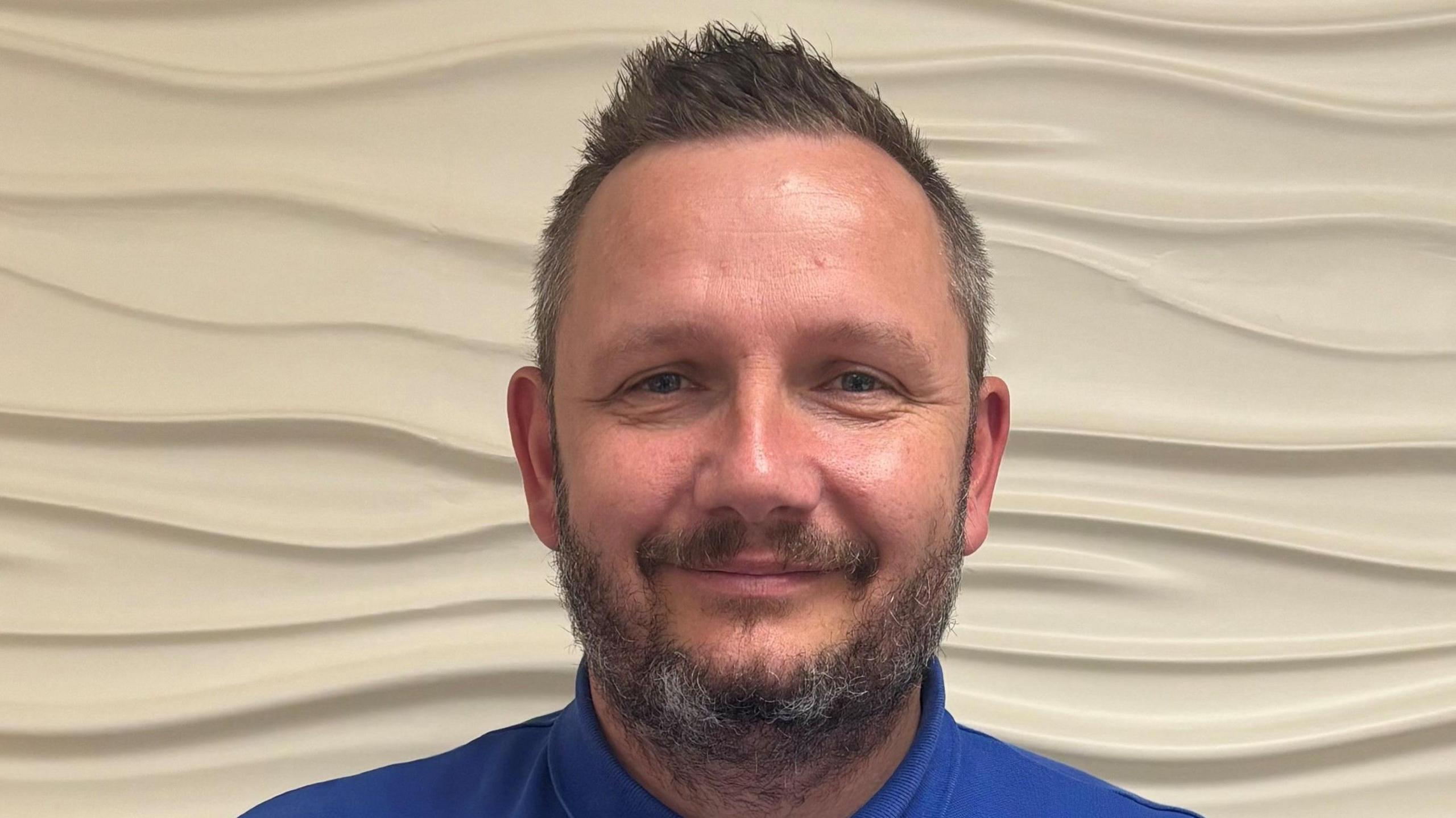 A profile image of Mark Curwood, an NHS worker wearing a blue polo shirt. He has brown, slightly greying hair. 
