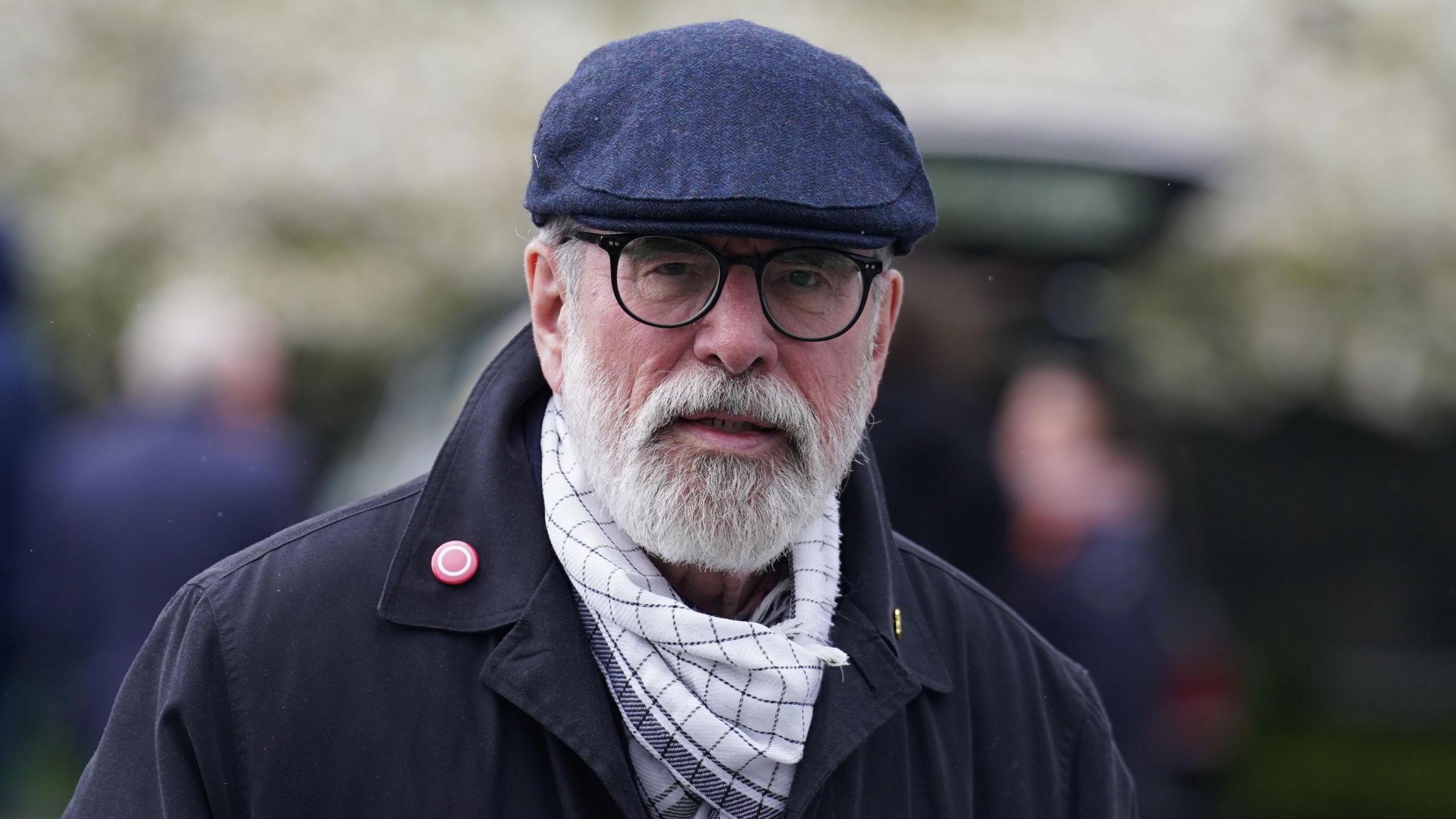 Gerry Adams wearing a dark coloured coat, a white scarf, a blue flat cap and black rimmed glasses. He has a grey beard. 