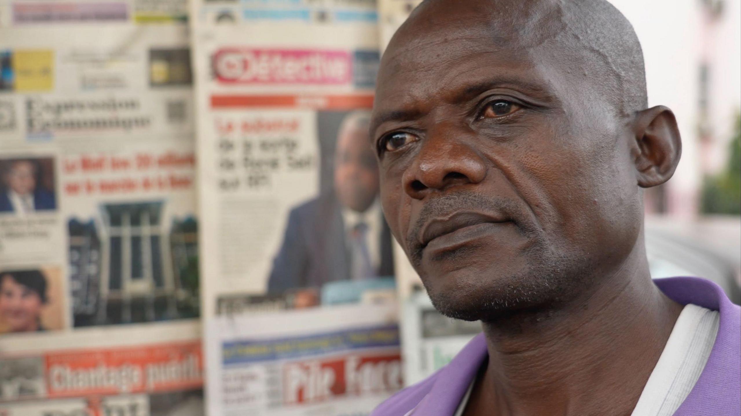 A close-up shot of Camille Esselem. He is wearing a purple shirt and has a clean-shaven head