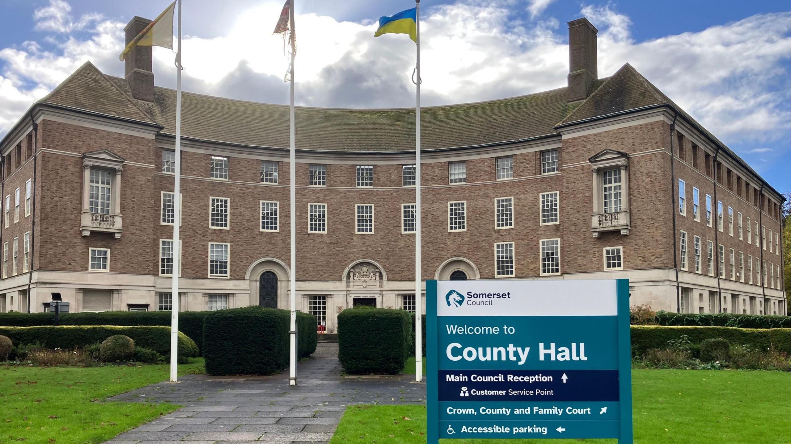 A municipal building, County Hall in Taunton, with a sign outside and three flags flying on a sunny day