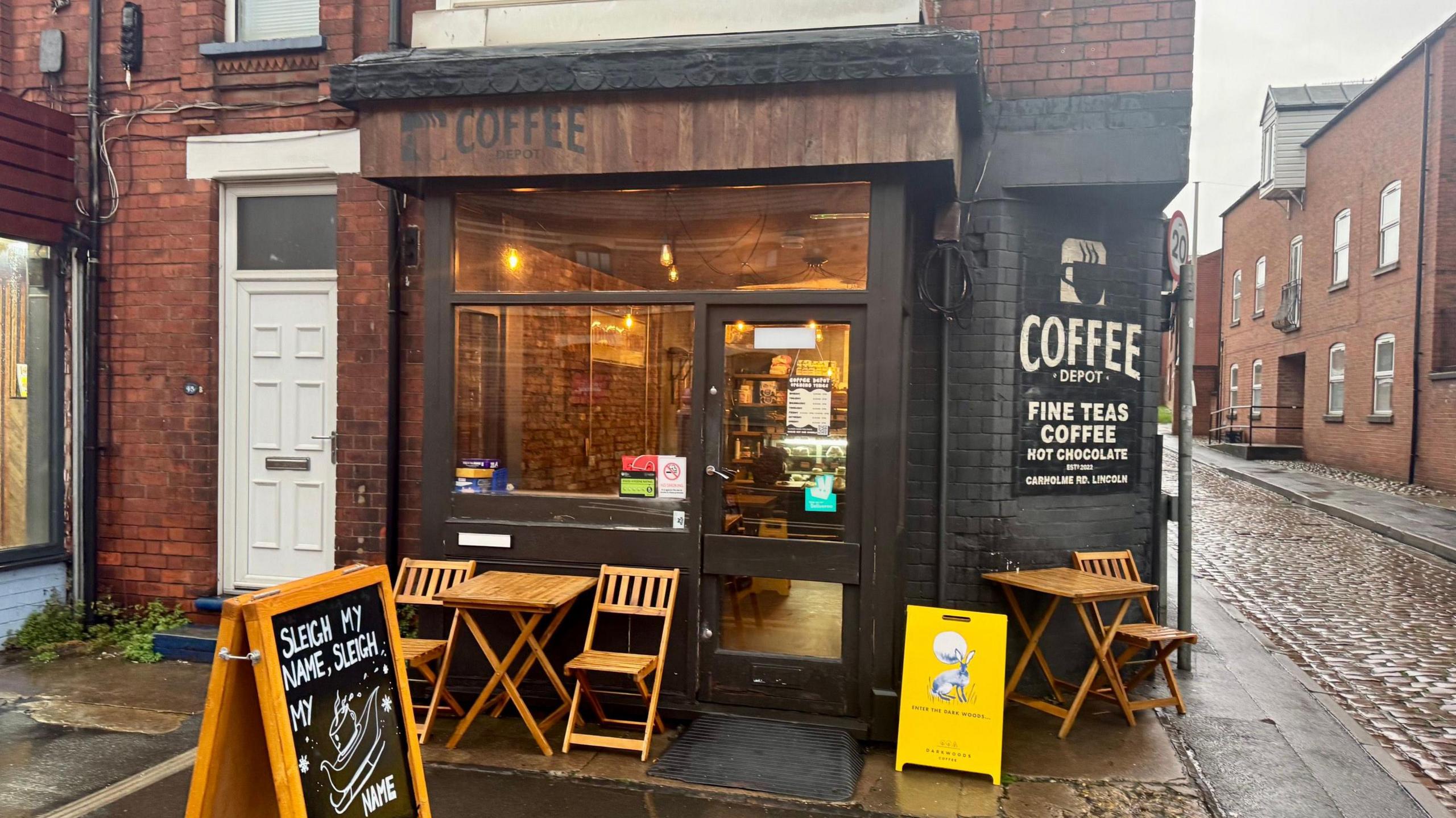 The front of a small coffee shop on the corner of a street. It has a dark wooden exterior with black writing 'Coffee Depot' written on it. It had black painted brick as well with more writing saying 'Coffee Depot, fine teas and coffee, hot chocolate'. A few wooden chairs and tables are placed outside with a sign in front saying 'Sleigh my name, sleigh my name'. The inside appears quiet with yellow lighting and brick wall decor.