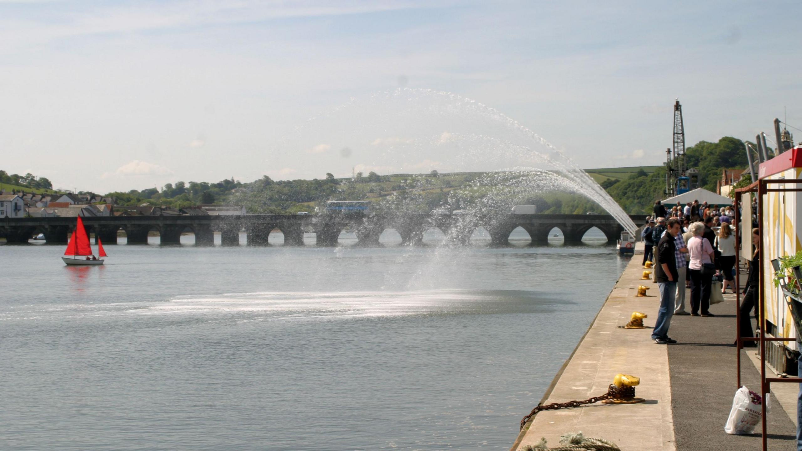 A fountain going into the river. There is a small sailing boat with red sails. There is a bridge going across the river. The sky is light blue. The water is a darker blue and the fountain water is clear. There are people walking along on the right.