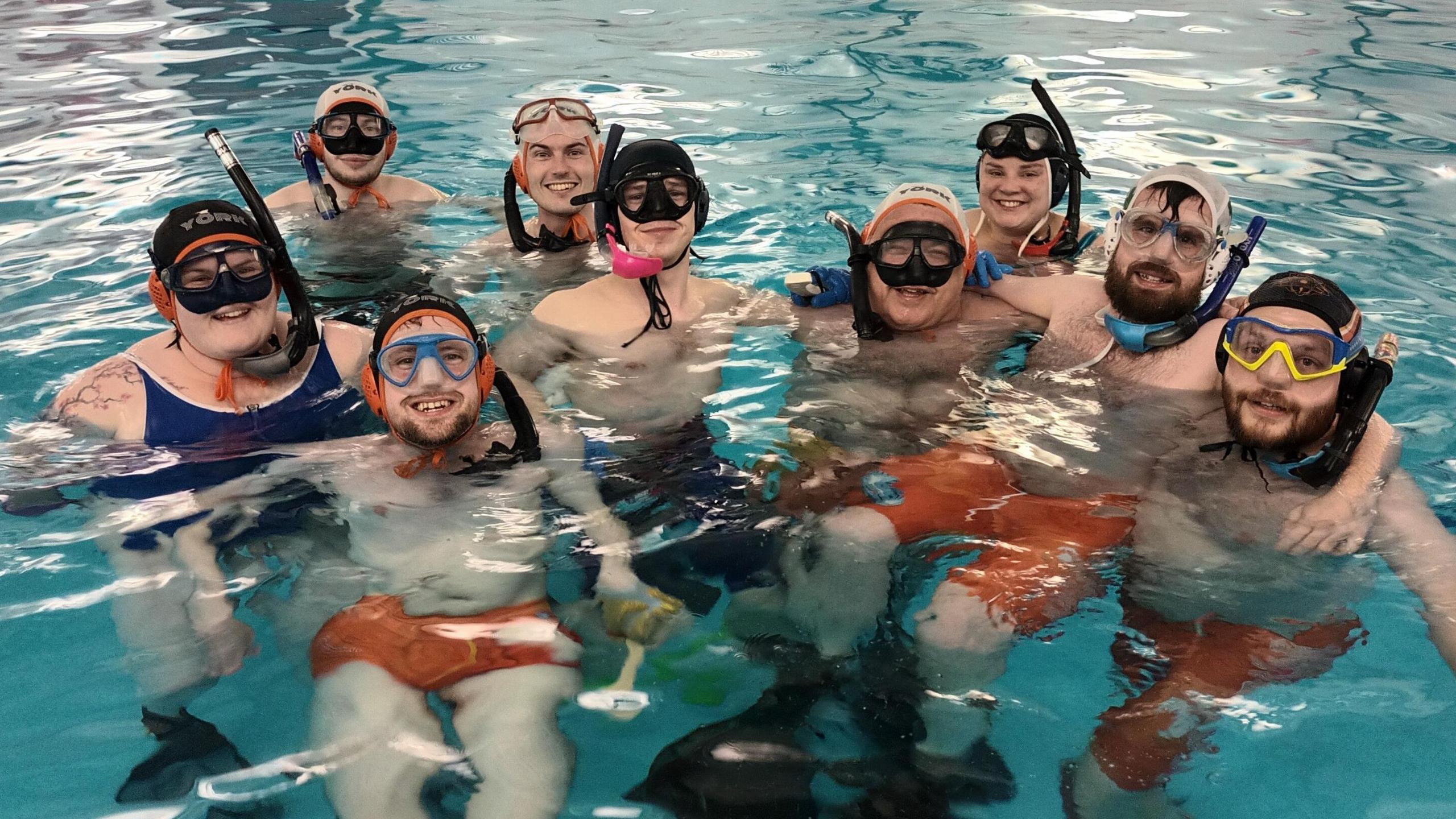 A mixed gender group of people pose with their arms around each other in a swimming pool. Everybody wears black snorkels, swimming caps and masks. The three people at the front can be seen wearing bright orange swimming trunks and flippers.