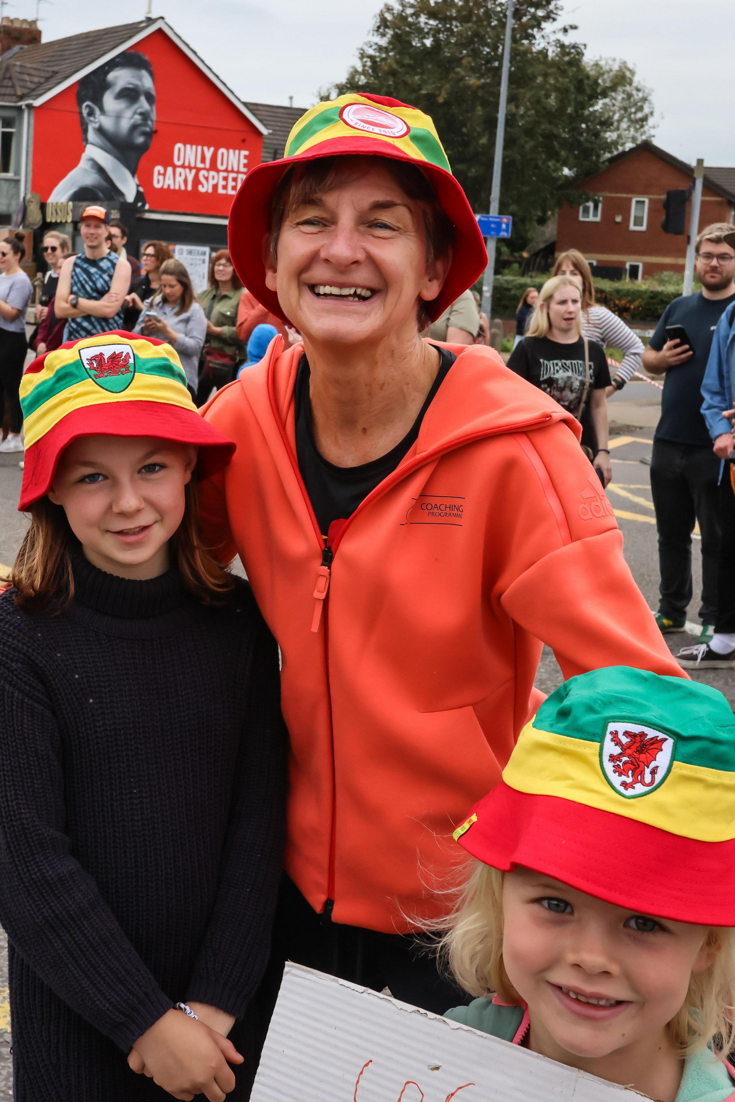 Laura McAllister wearing a bucket hat standing with two children also wearing bucket hats. There are crowds behind her cheering as runners go past from the Cardiff Half Marathon with a red mural of Gary Speed seen on a wall to the right side of the image.