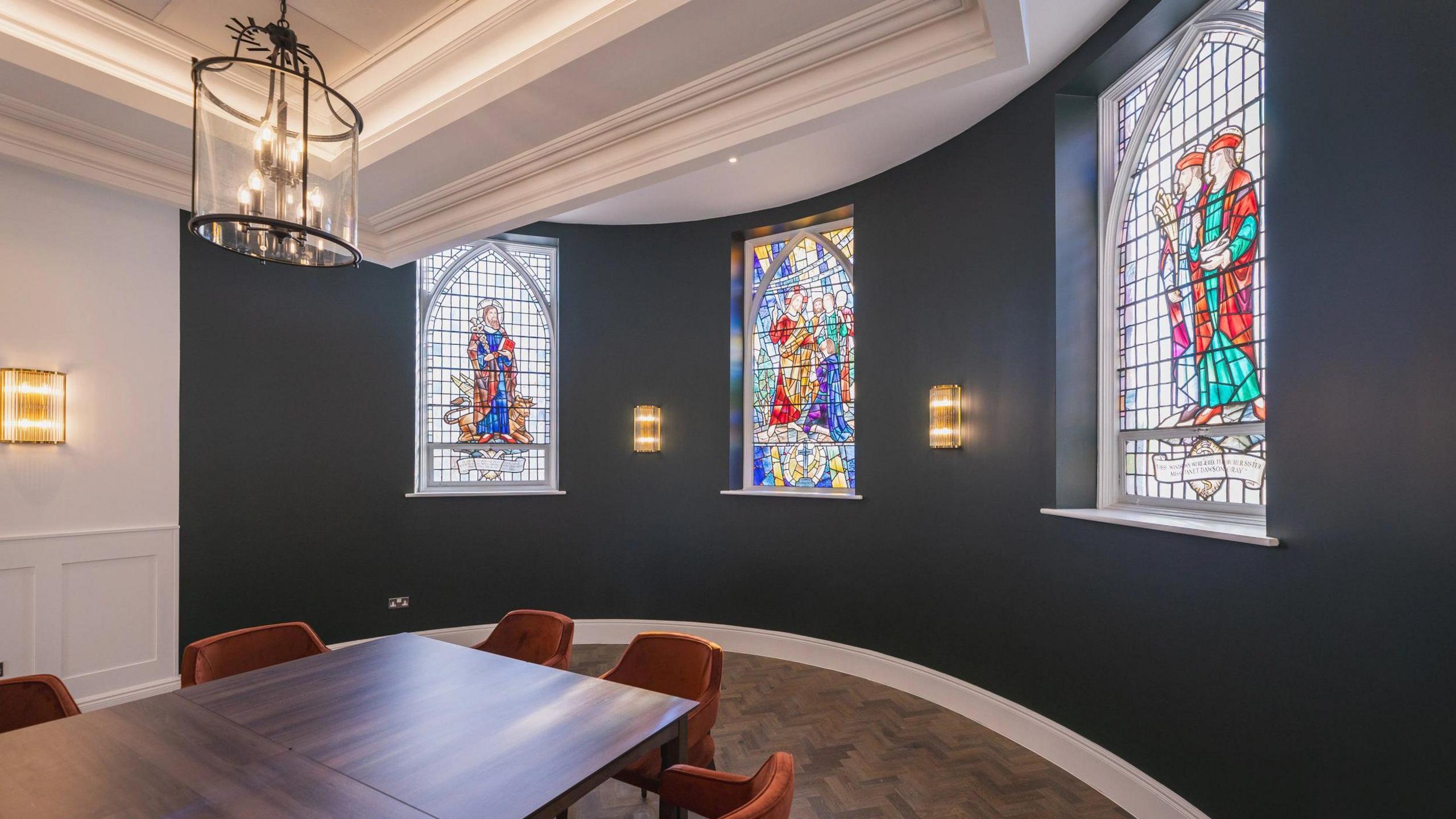 An empty room with a modern table and chairs, decorated in dark grey and white, but with the hospital's old stained glass still there.