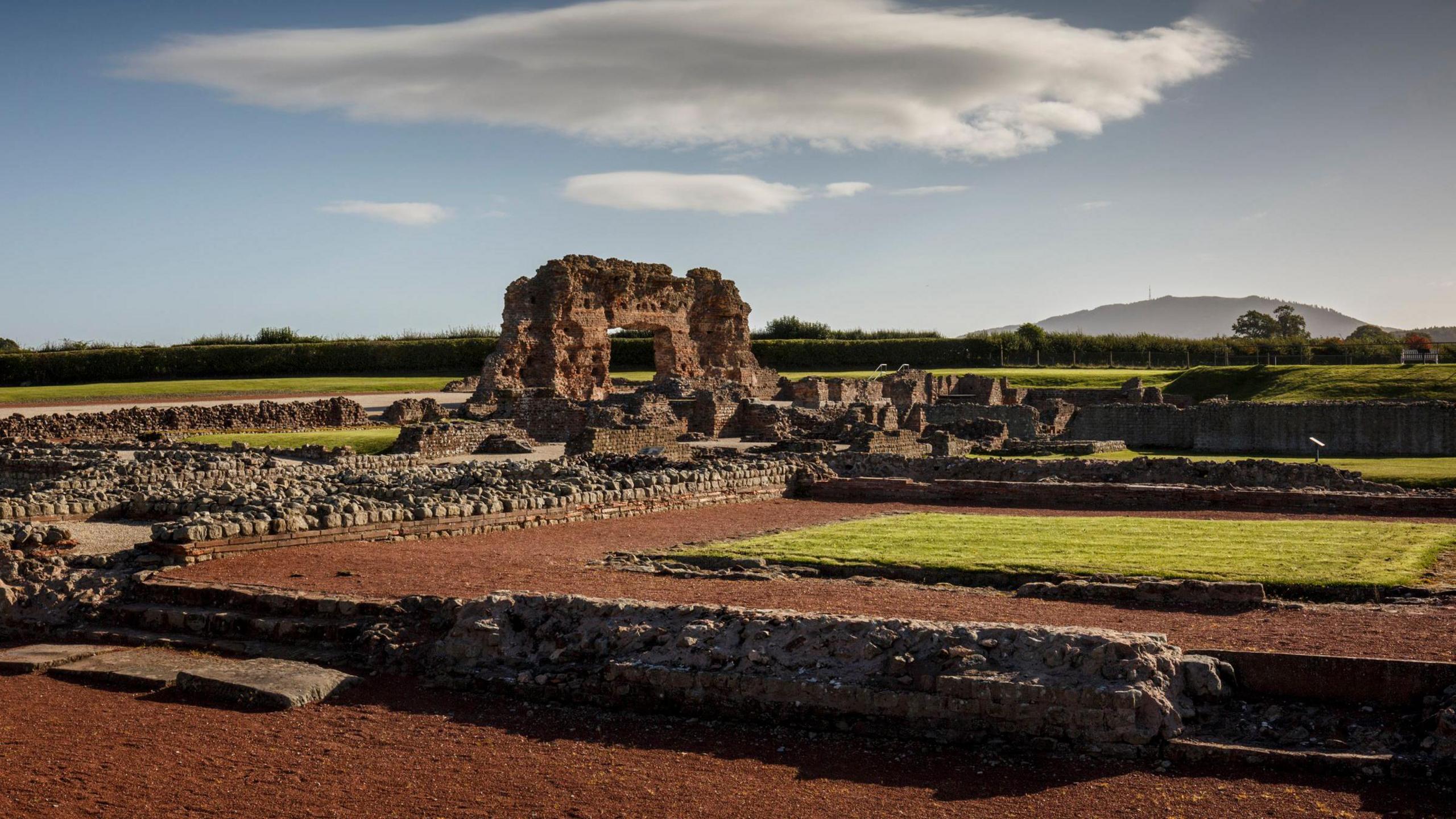 Wroxeter Roman City. The picture shows brown earth with short stone walls which once formed buildings. There is a brown stone archway standing and surrounded by green grass.