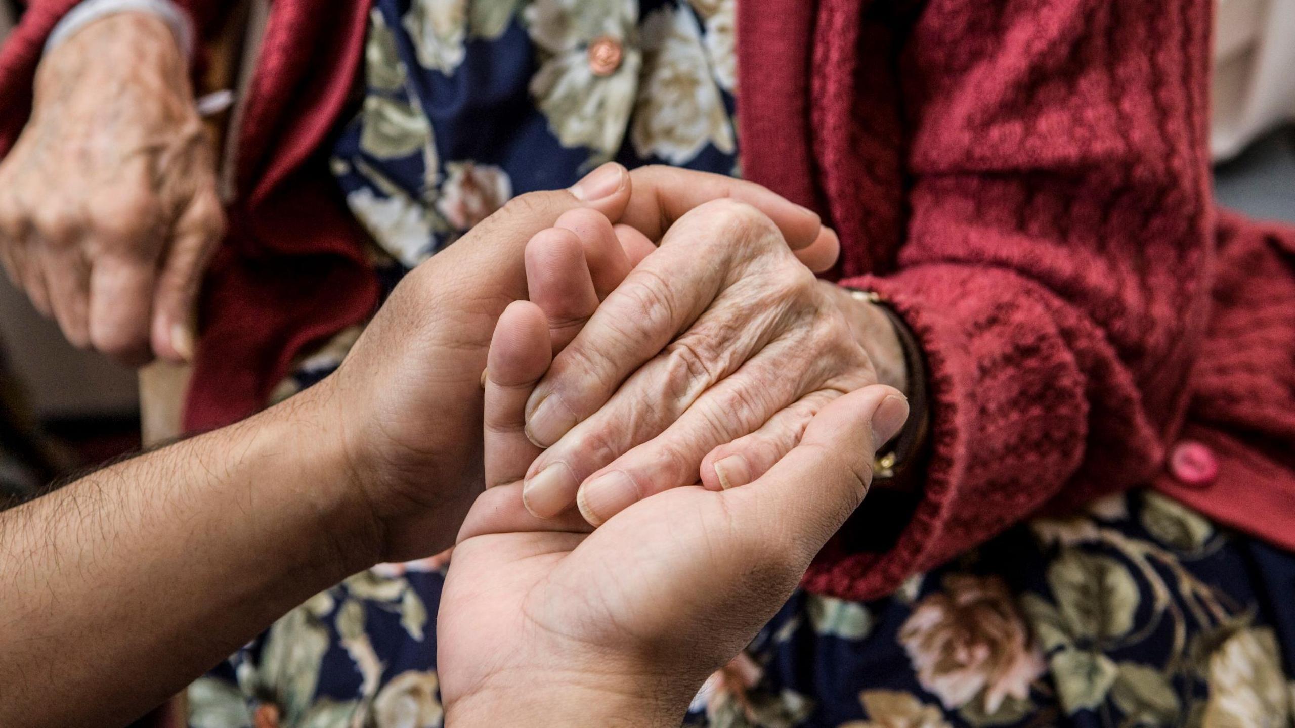 An old person in a red knitted cardigan and a flora dress having her hand held by another person