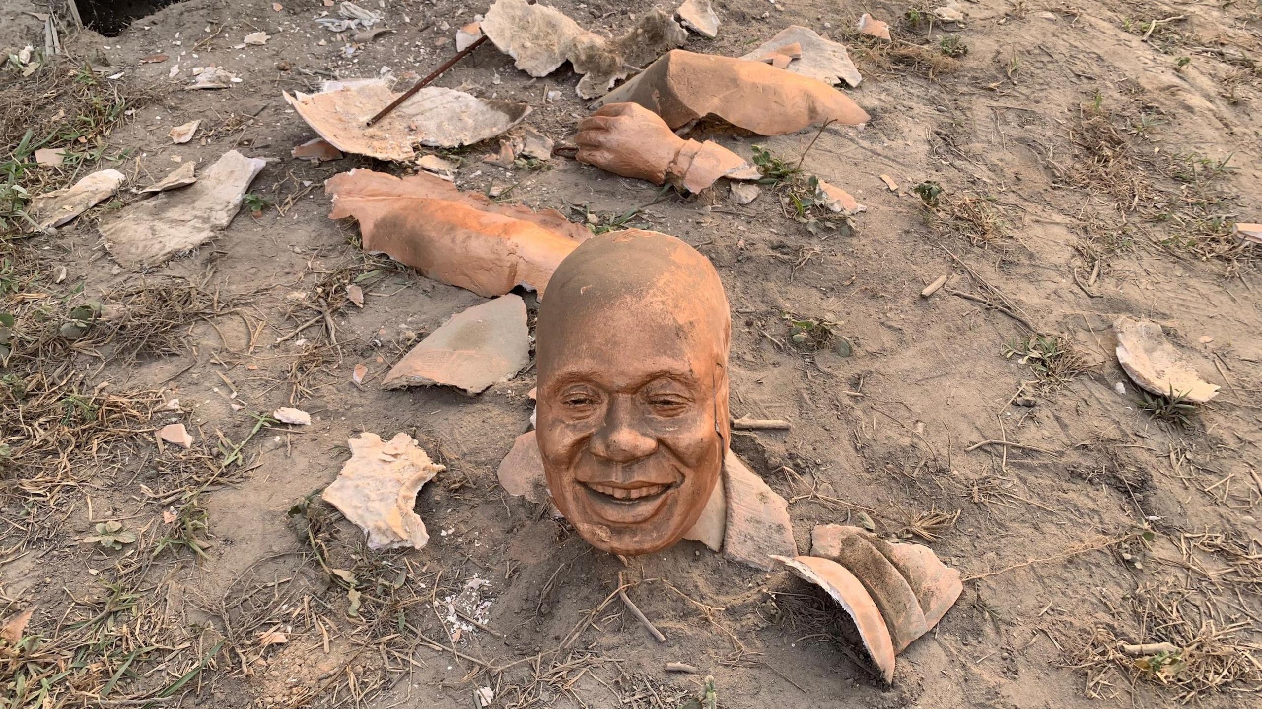 The severed head of from the statue of Nana Akufo-Addo, bronze in colour, lying on the ground. There are fragments of other parts of the statue on the dirt ground