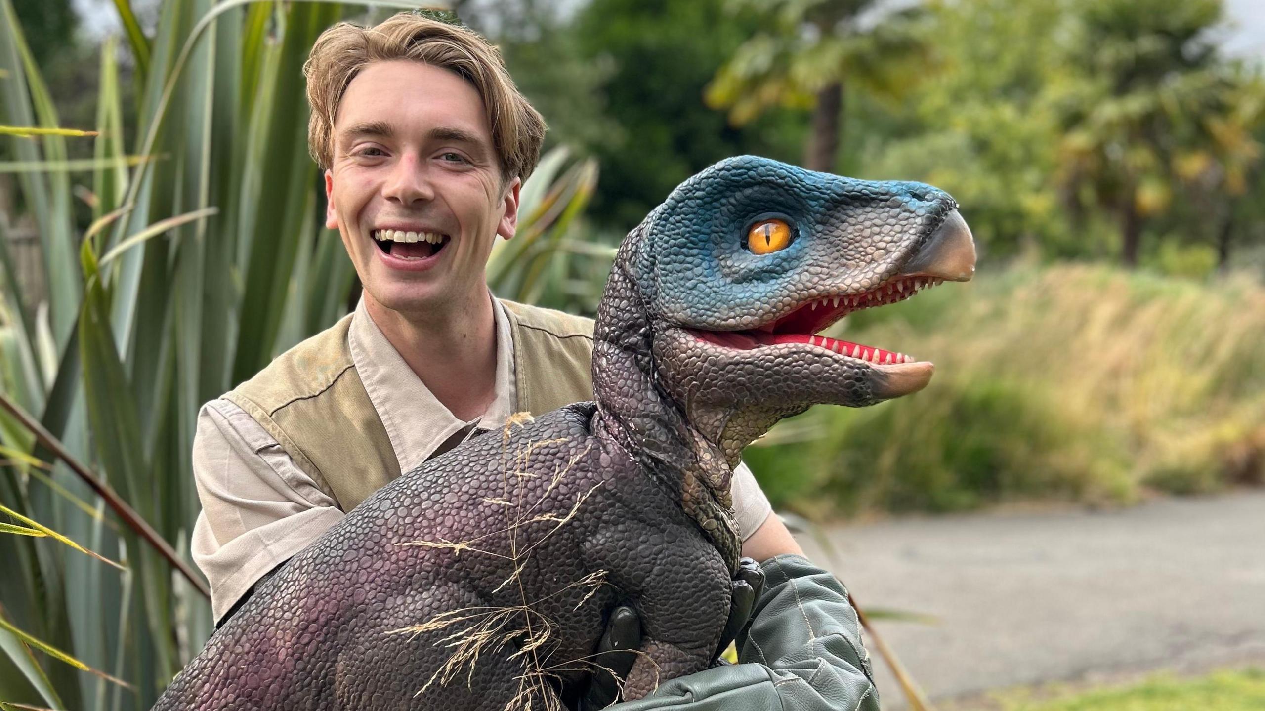 A man, wearing a off white top and light green vest, smiling at the camera holding a velociraptor. The velociraptor is on it's side, it has purple scales on it's body and blue scales on its head and has it's mouth open and a big orange eye. Behind them is green bushes and trees and a concrete path, the background is blurred.