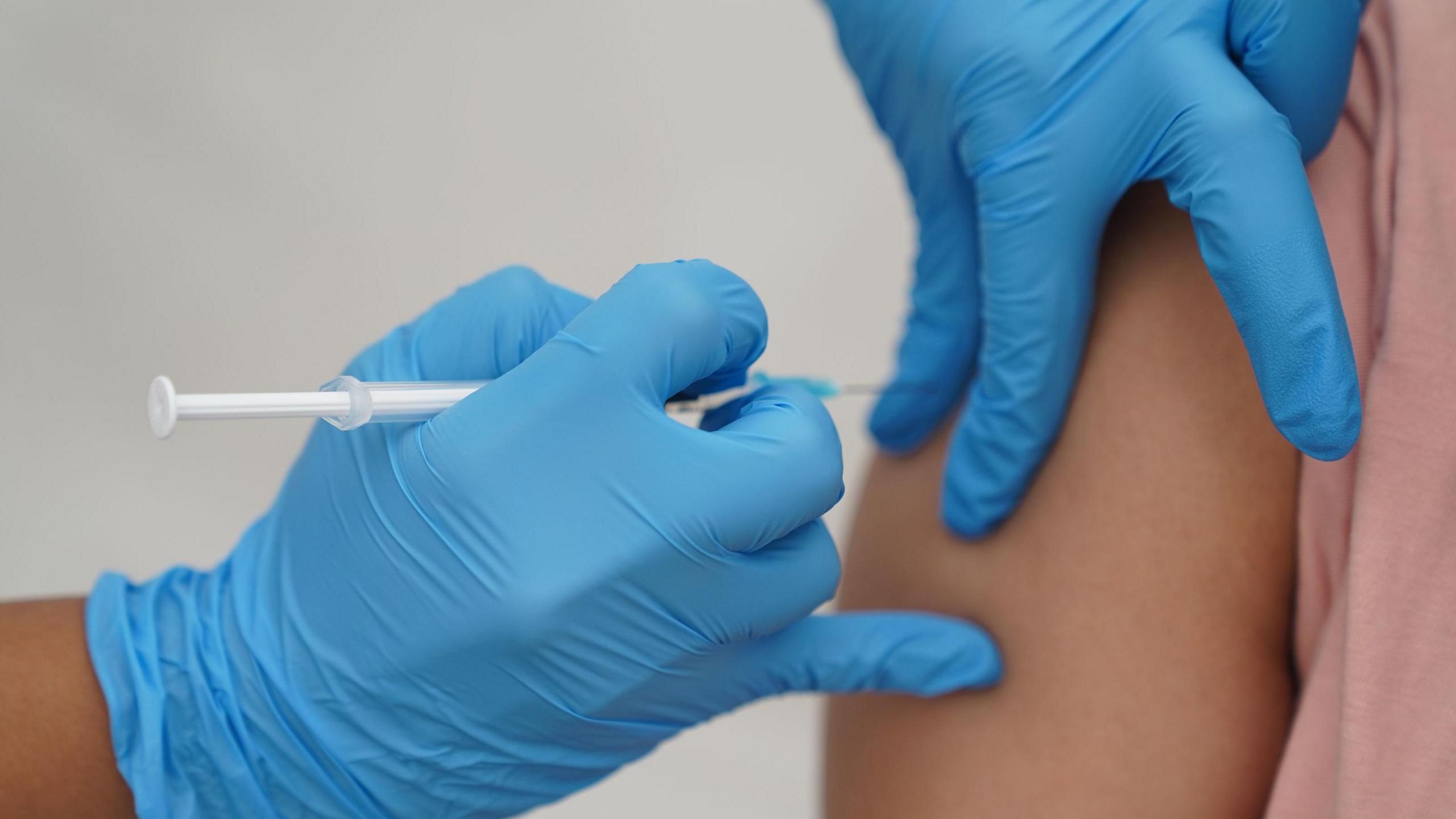 A nurse wearing blue medical gloves injects a vaccination into a patient's upper arm.