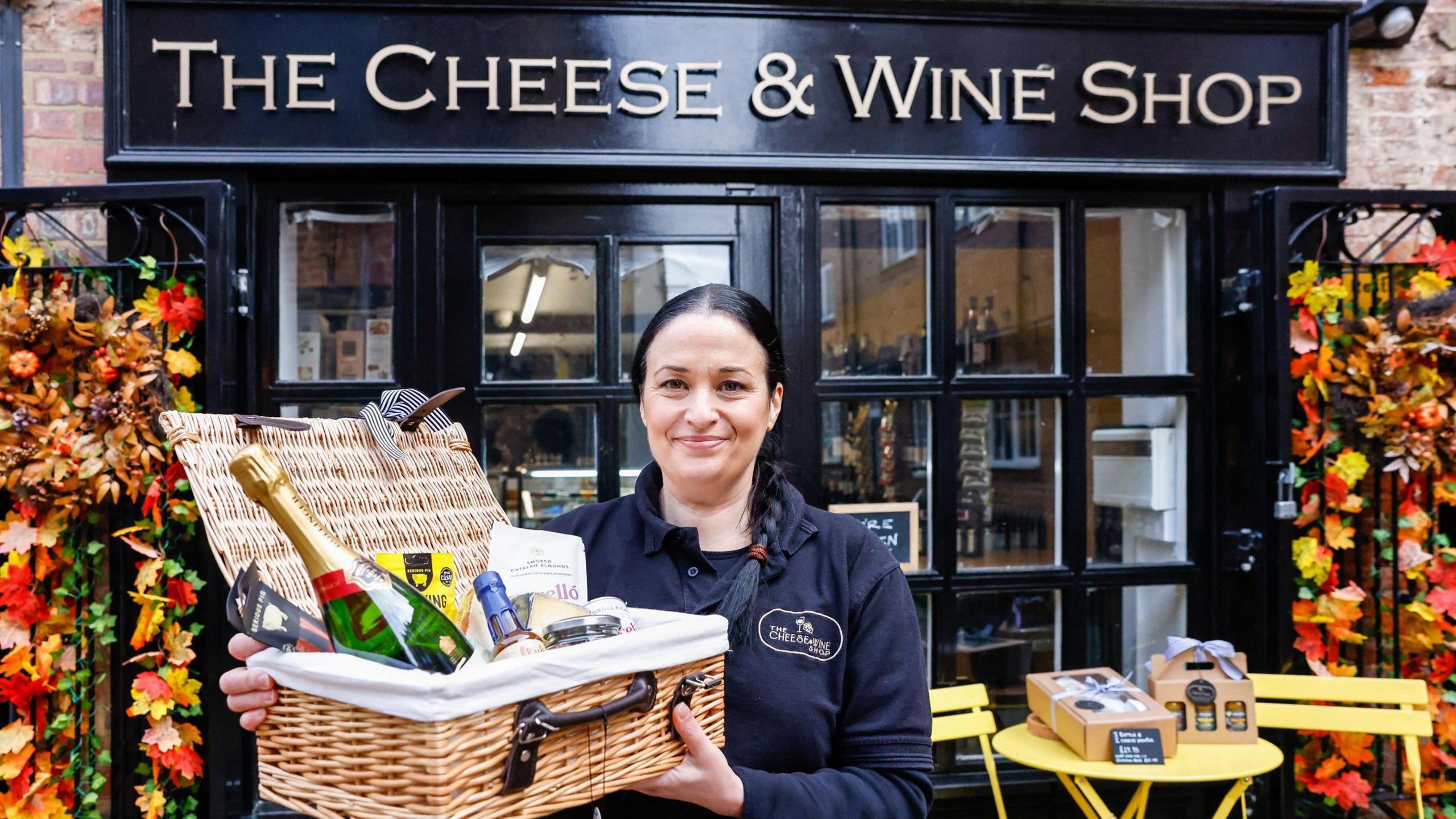 Caroline Elliott standing outside The Cheese & Wine Shop holding a wicker basket of food and drink. She has black hair tied into a pony tail and is wearing a navy top. The shopfront is navy with gold writing above the door and windows. A yellow table with two matching chairs is outside.