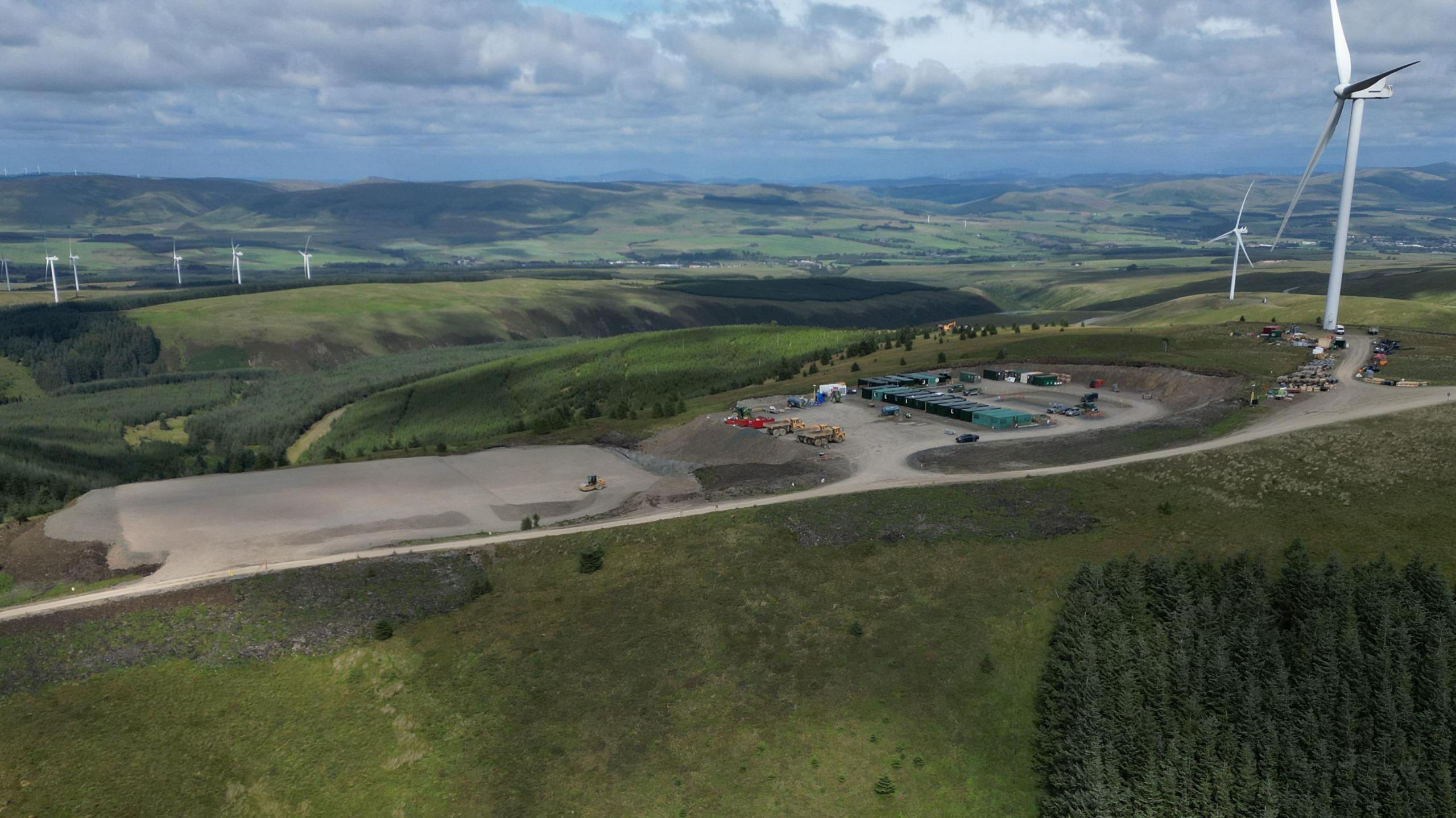 Drone shot looking down on a construction site. A number of diggers, other vehicles and temporary buildings are on site. There are some large, white wind turbines in an area surrounded by fields and trees 