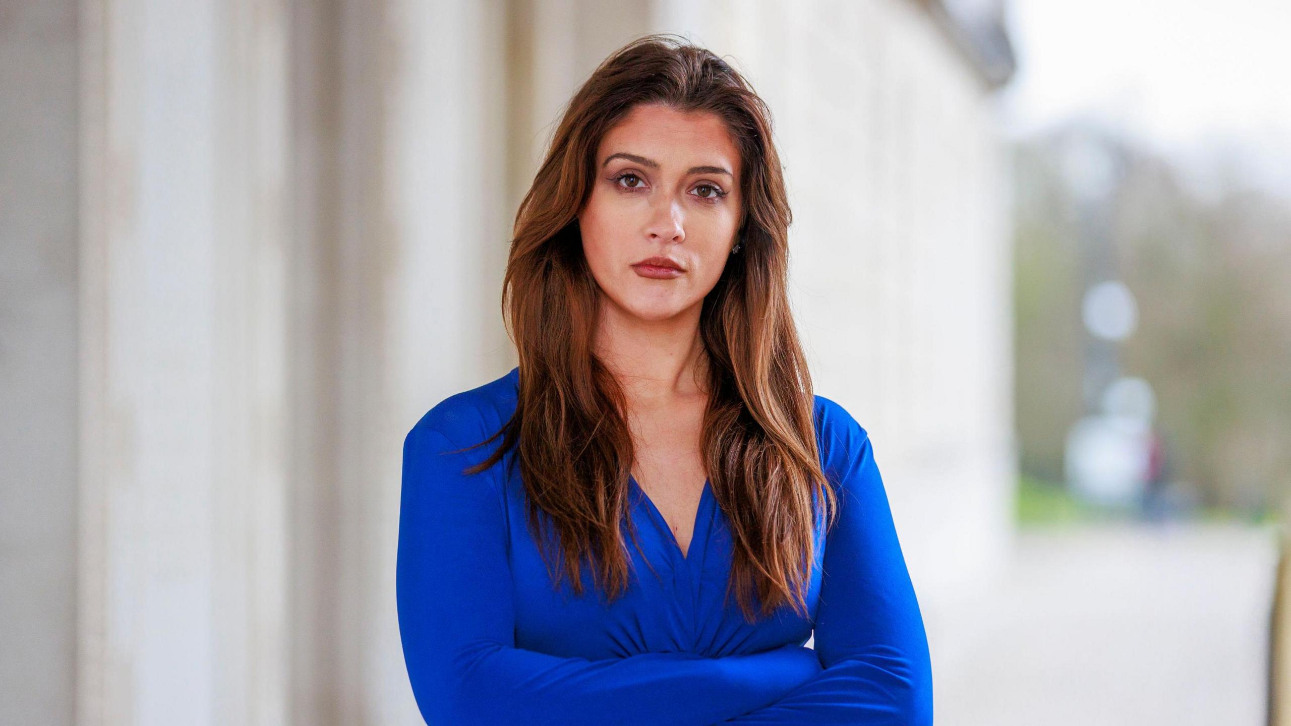 Cara Hunter standing outside Stormont with her arms folded.   She has long, dark hair and is wearing a blue dress.  She is looking at the camera.