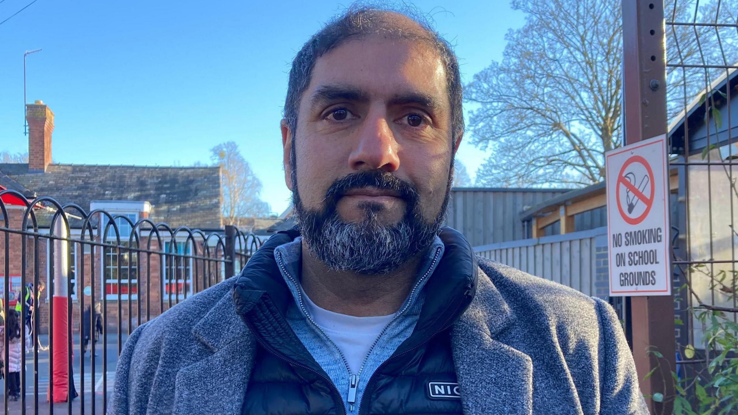 Jabba Riaz standing outside a school, wearing a blue shirt and grey coat