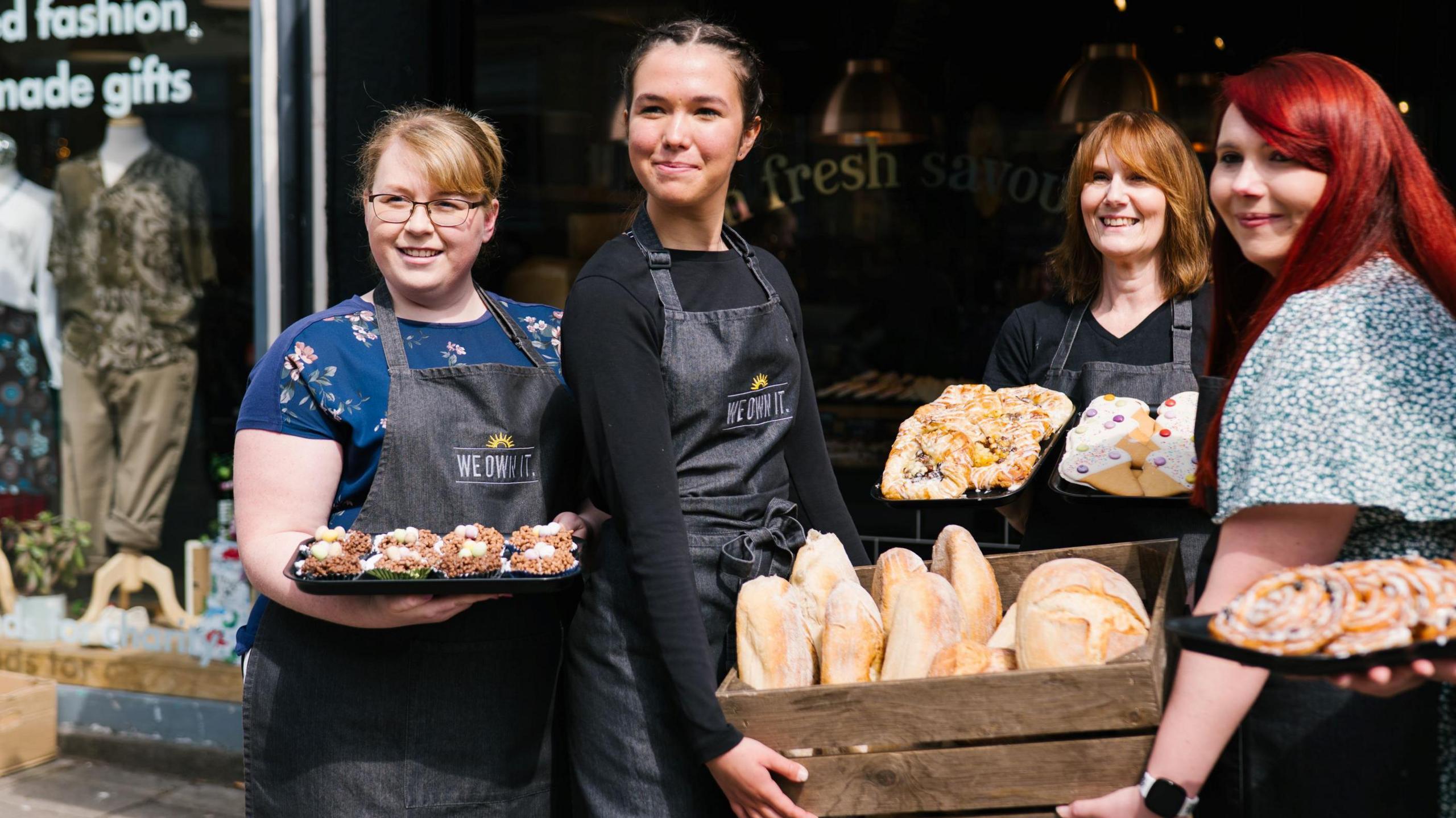 Four female employees of Bristol bakery Parsons stand and smile at the camera, while also holding bread products and cakes