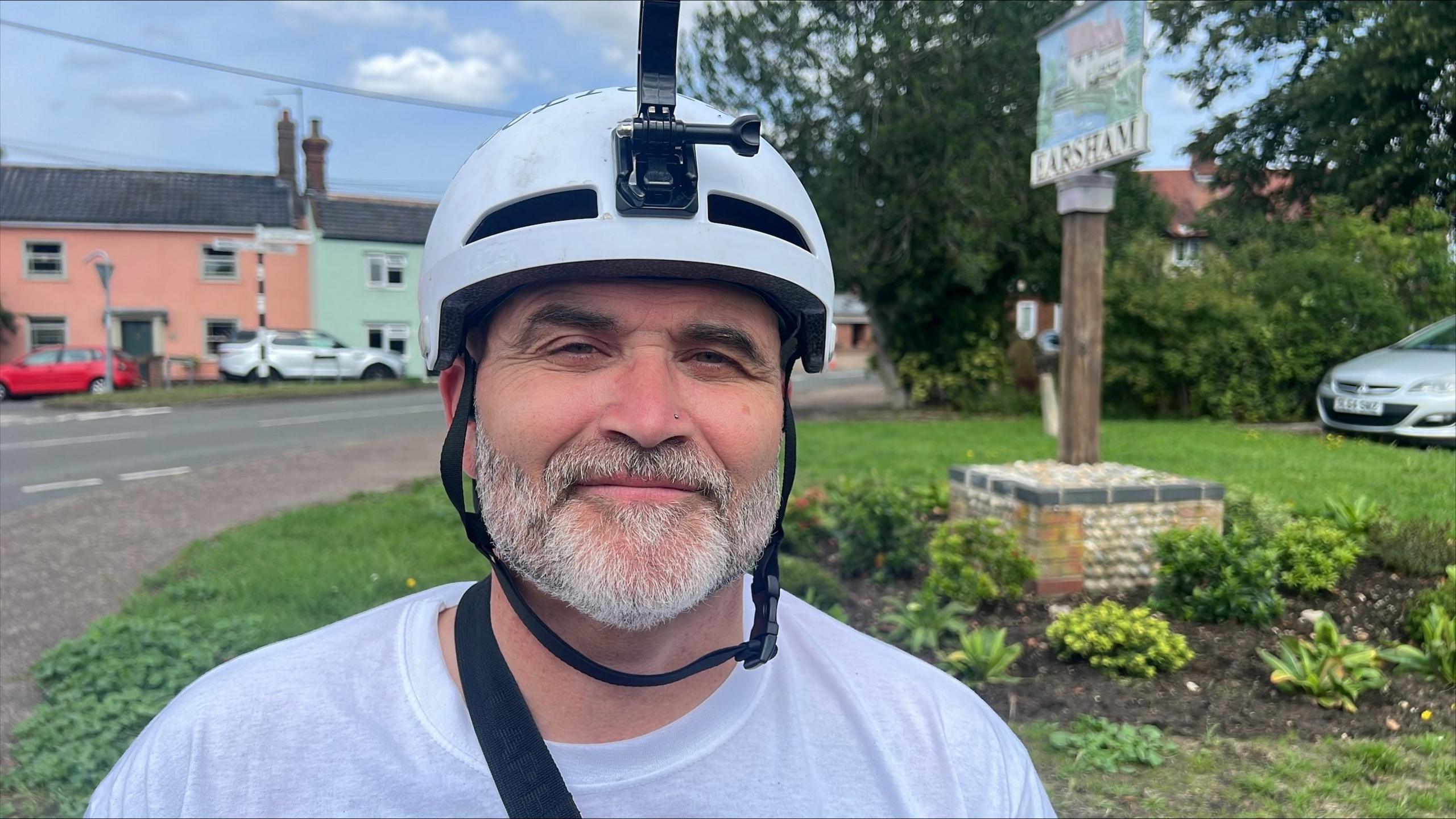 Ben Parker smiles at the camera. He wears a white helmet with a camera attached to it and a white T-shirt. A village centre can be seen behind him.