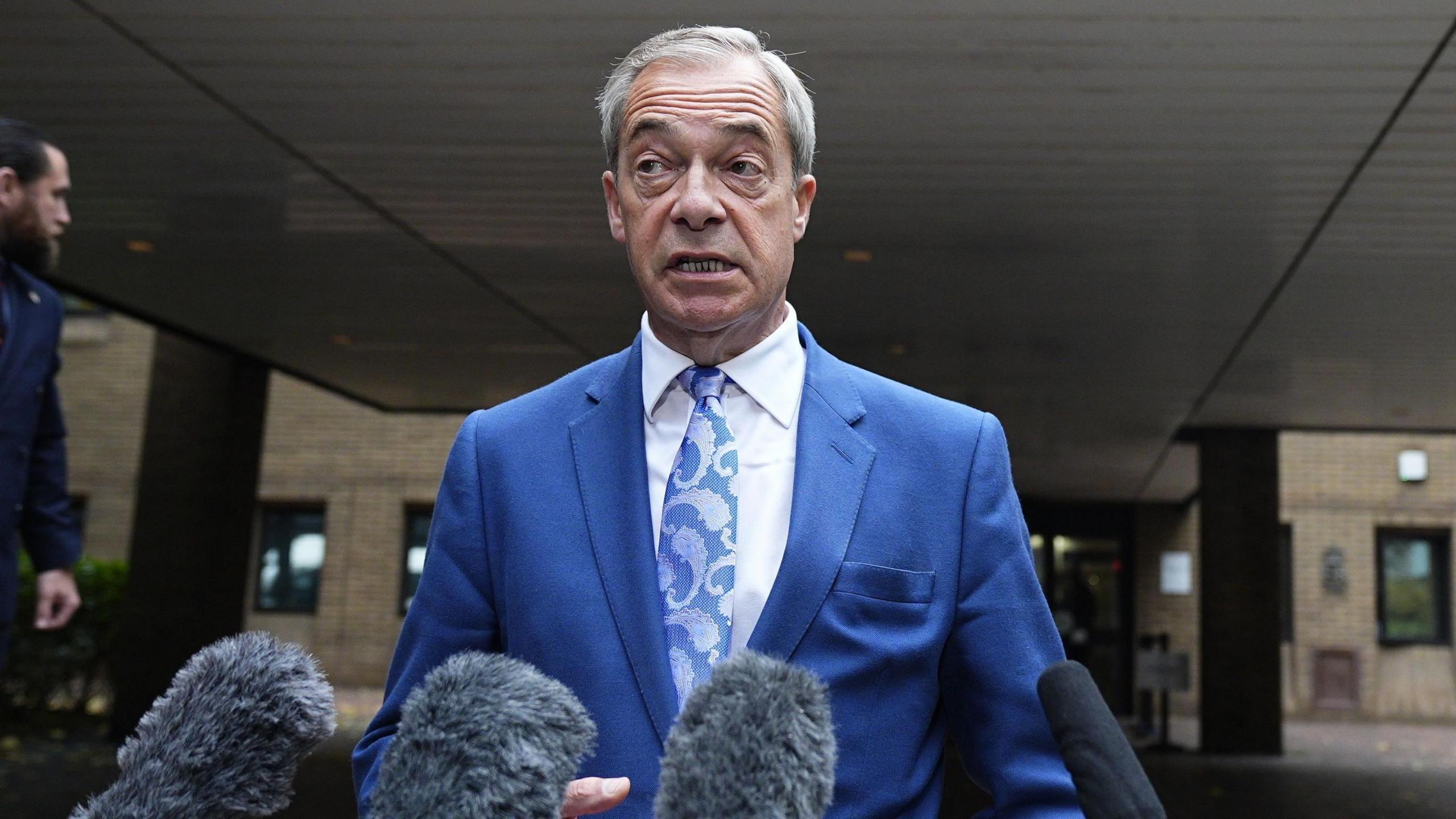 Nigel Farage on the court steps speaking to the gathered press. He has microphones in front of him and is wearing a blue suit jacket, blue and silver tie and a white shirt.