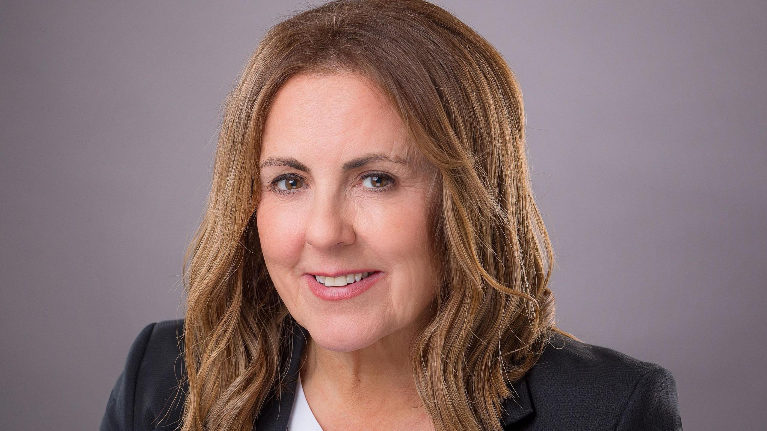 A woman with long, dark blonde hair and wearing a suit jacket and white top, smiles while in front of a purple background.
